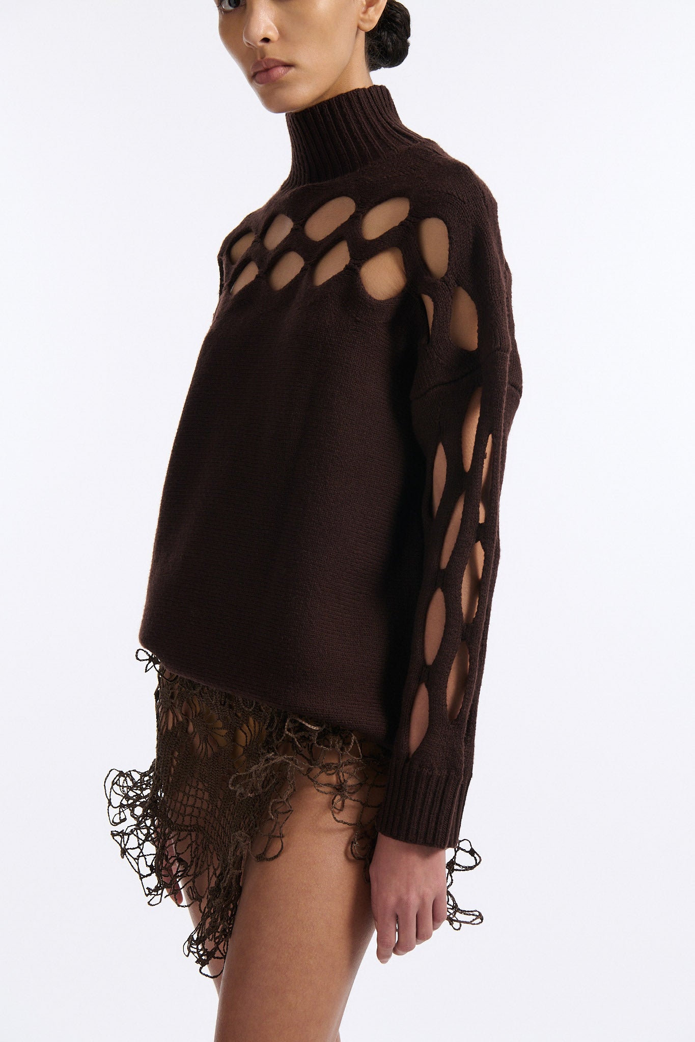 Female model with slicked back black hair in a bun wears brown knit mockneck with horizontal diamond cutouts along arms, chest, and back. Model also wears a see-through crochet mini skirt in brown and white wedges.