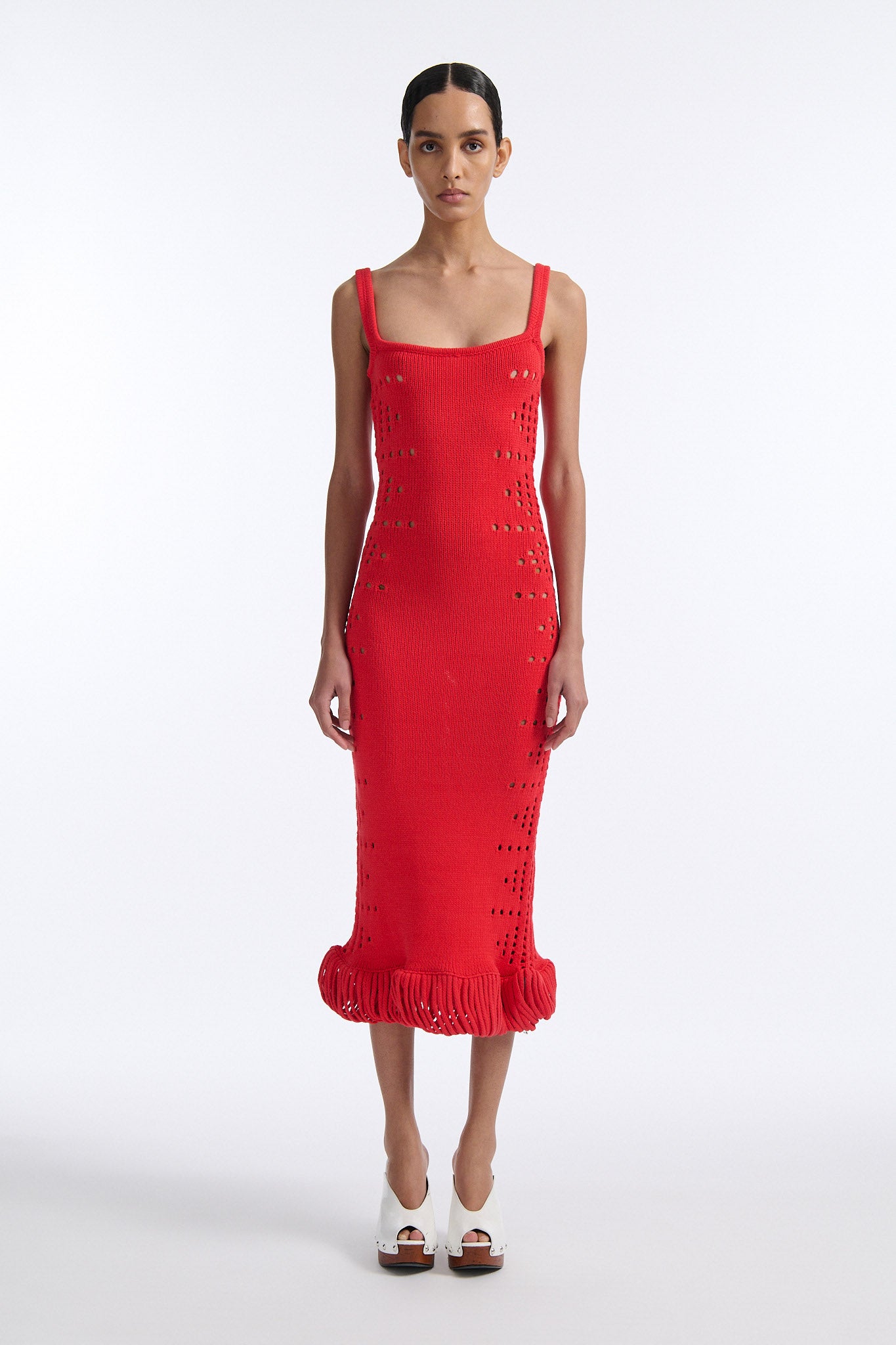 Female model with slicked back black hair in a bun wears a hibiscus red camisole knit dress. Dress is long with oversized pointelle detailing along both sides and looped fringe hem.