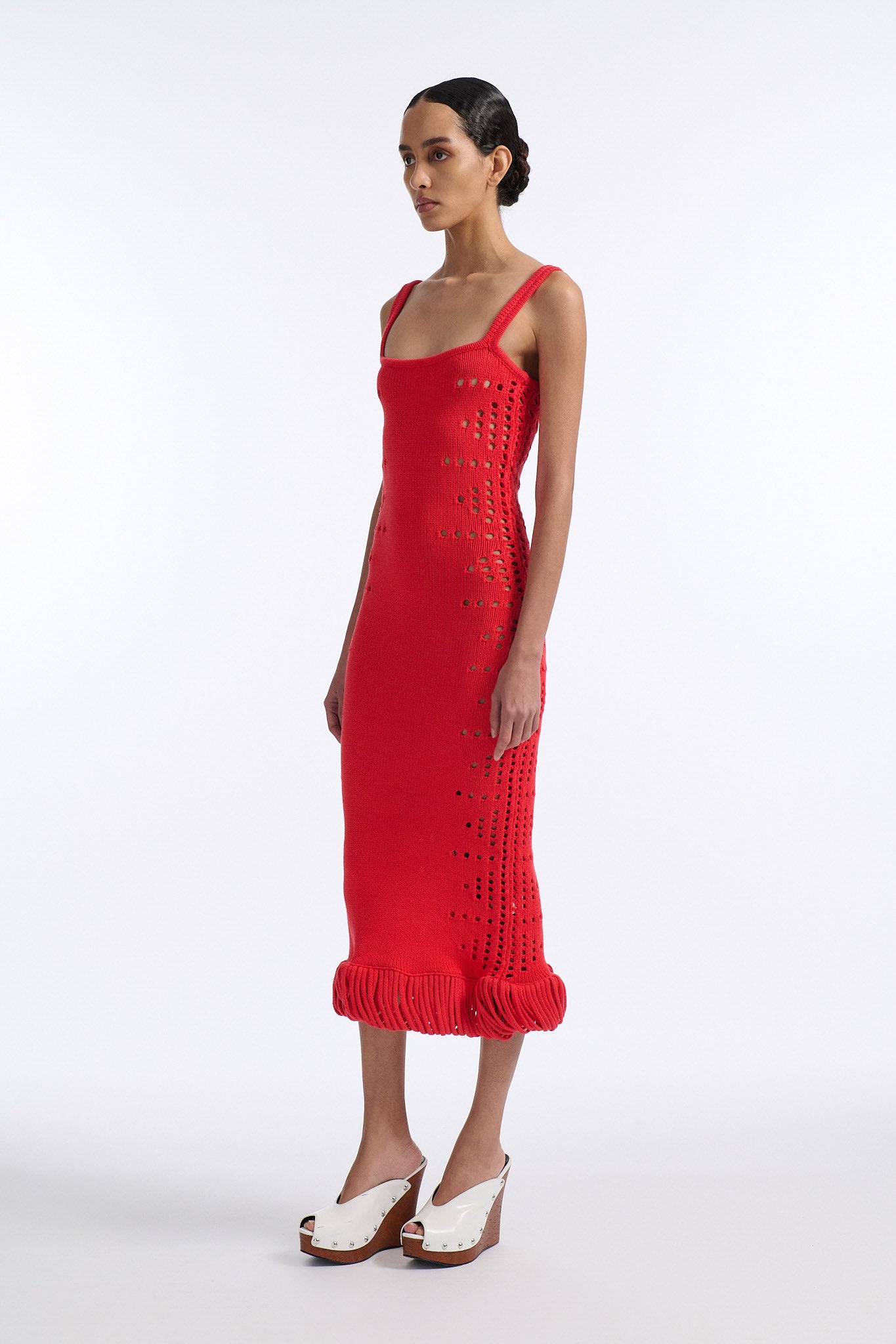 Female model with slicked back black hair in a bun wears a hibiscus red camisole knit dress. Dress is long with oversized pointelle detailing along both sides and looped fringe hem.