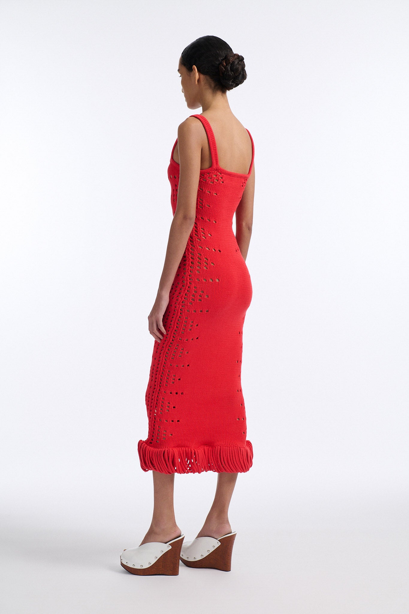 Female model with slicked back black hair in a bun wears a hibiscus red camisole knit dress. Dress is long with oversized pointelle detailing along both sides and looped fringe hem.