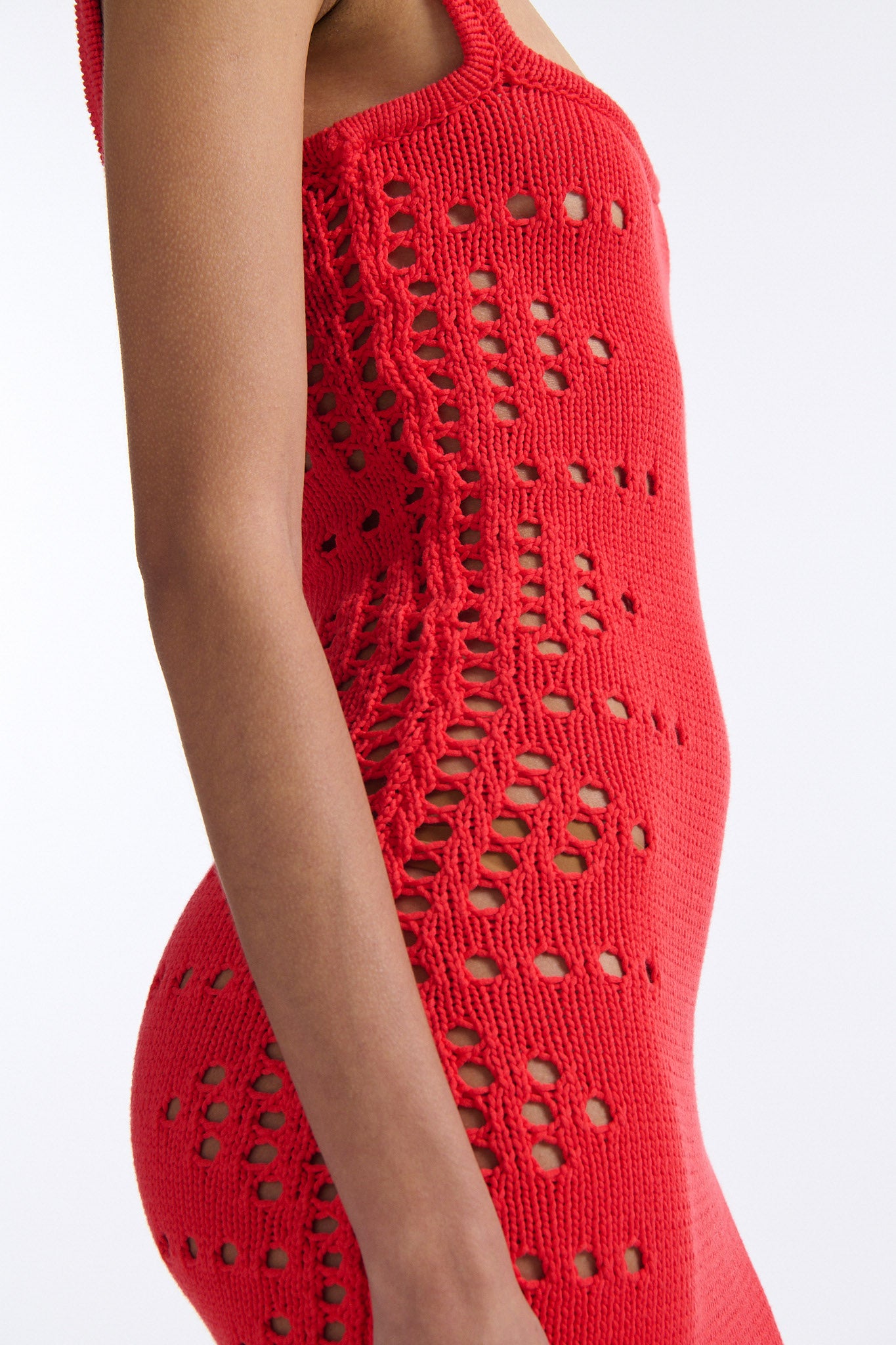 Female model with slicked back black hair in a bun wears a hibiscus red camisole knit dress. Dress is long with oversized pointelle detailing along both sides and looped fringe hem.