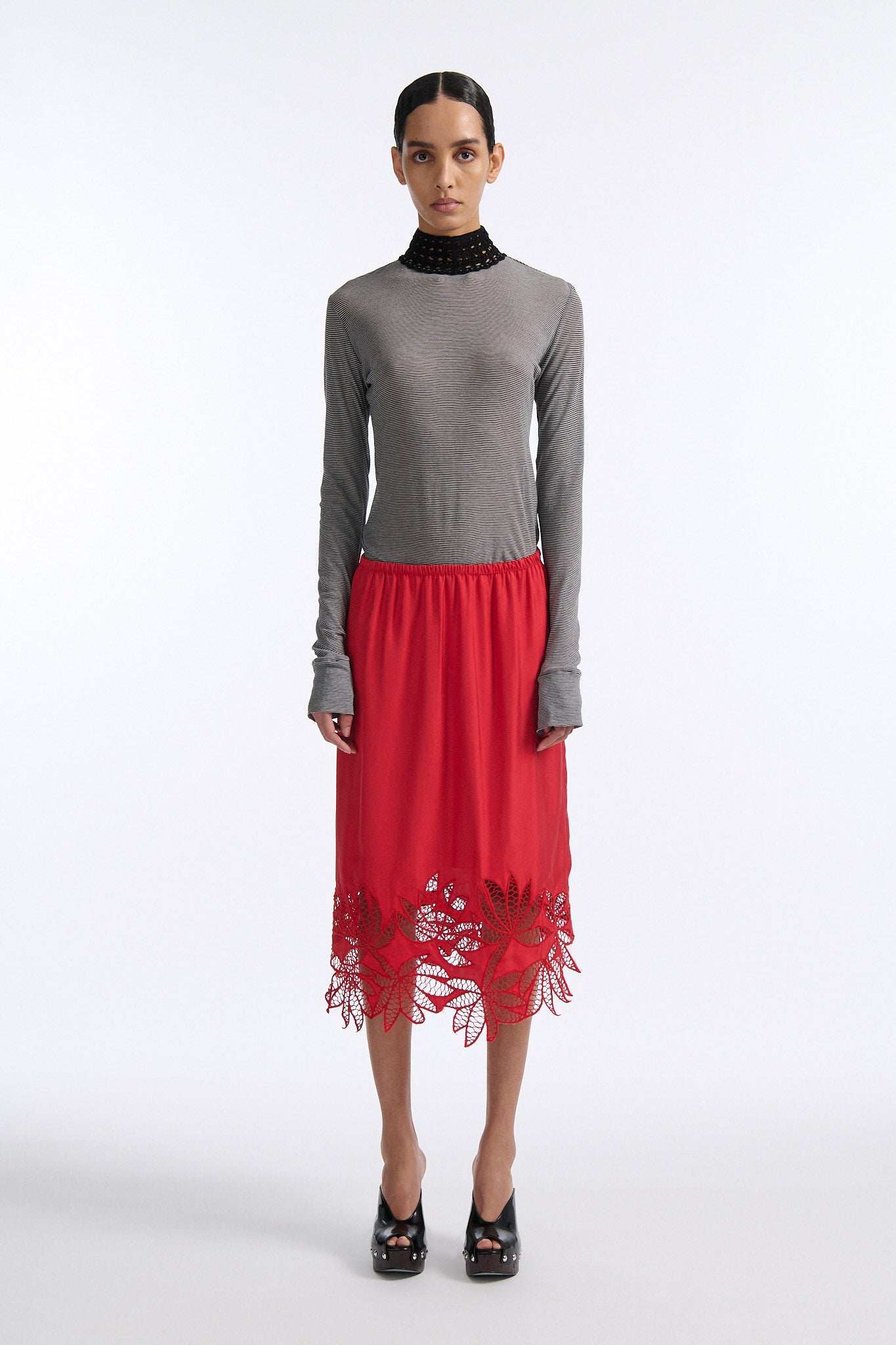 Female model with slicked back black hair in a bun wears black and grey striped fine-gauge cotton jersey long sleeve top with black mockneck crochet collar. Model also wears red silk shift skirt with palm leaf cutouts at hem. Skirt has elastic waist and is mid-length.