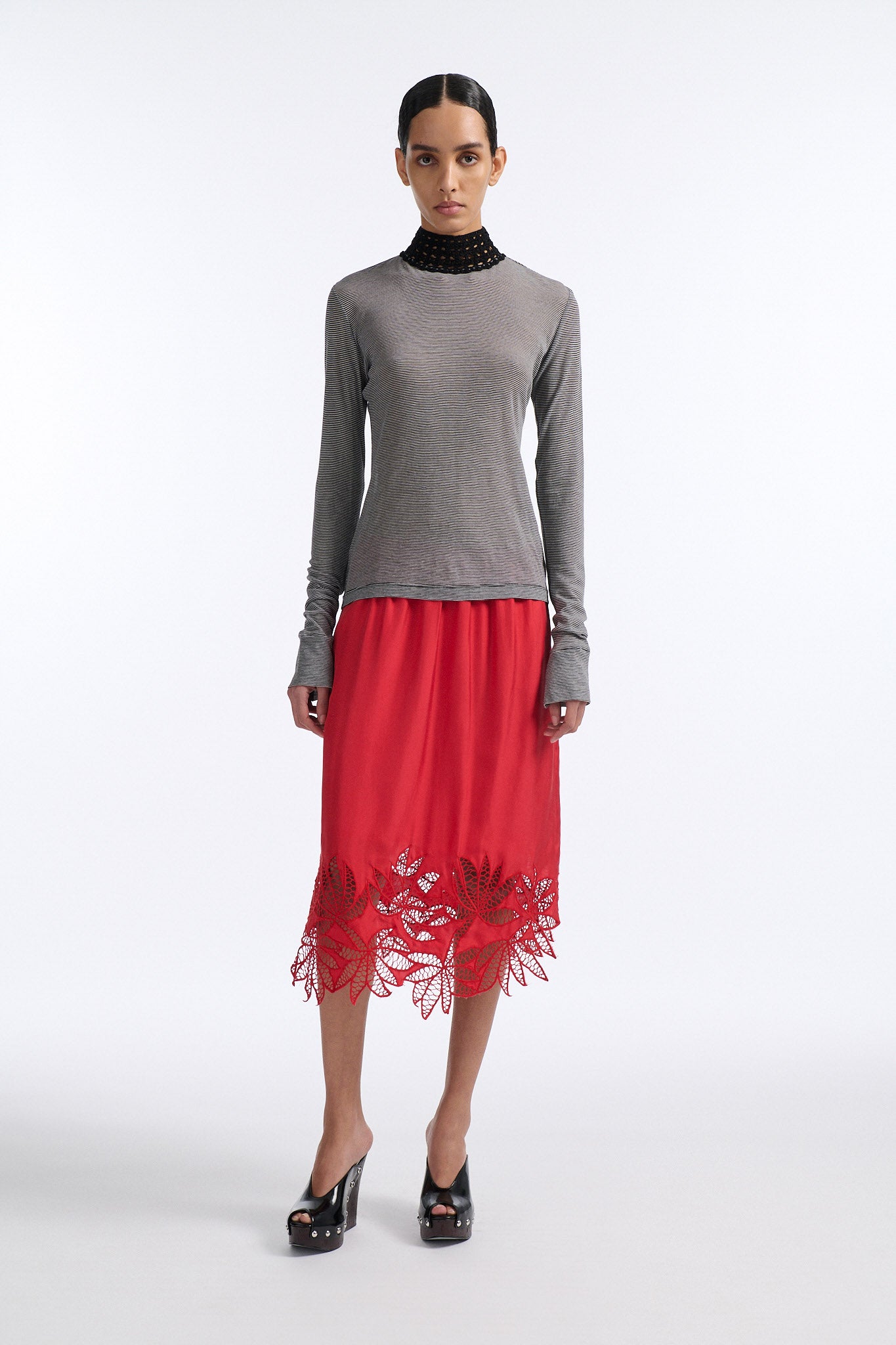 Female model with slicked back black hair in a bun wears black and grey striped fine-gauge cotton jersey long sleeve top with black mockneck crochet collar. Model also wears red silk shift skirt with palm leaf cutouts at hem and elastic waist.