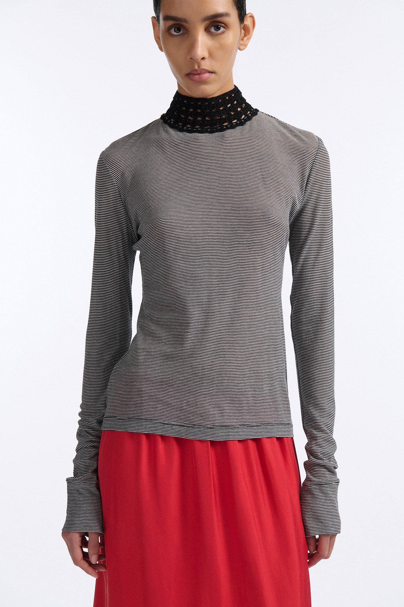 Female model with slicked back black hair in a bun wears black and grey striped fine-gauge cotton jersey long sleeve top with black mockneck crochet collar. Model also wears red silk shift skirt with palm leaf cutouts at hem and elastic waist.