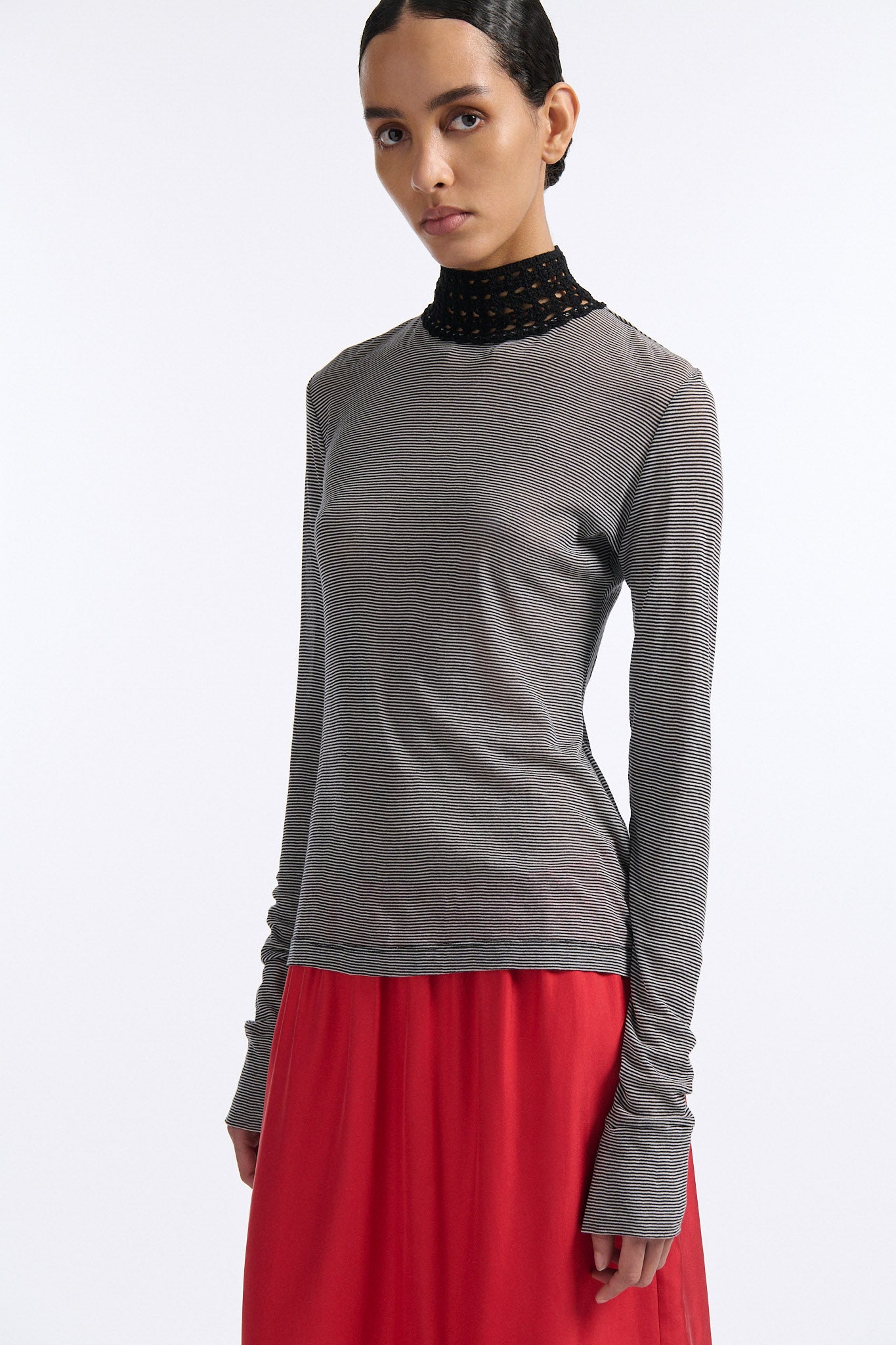 Female model with slicked back black hair in a bun wears black and grey striped fine-gauge cotton jersey long sleeve top with black mockneck crochet collar. Model also wears red silk shift skirt with palm leaf cutouts at hem and elastic waist.