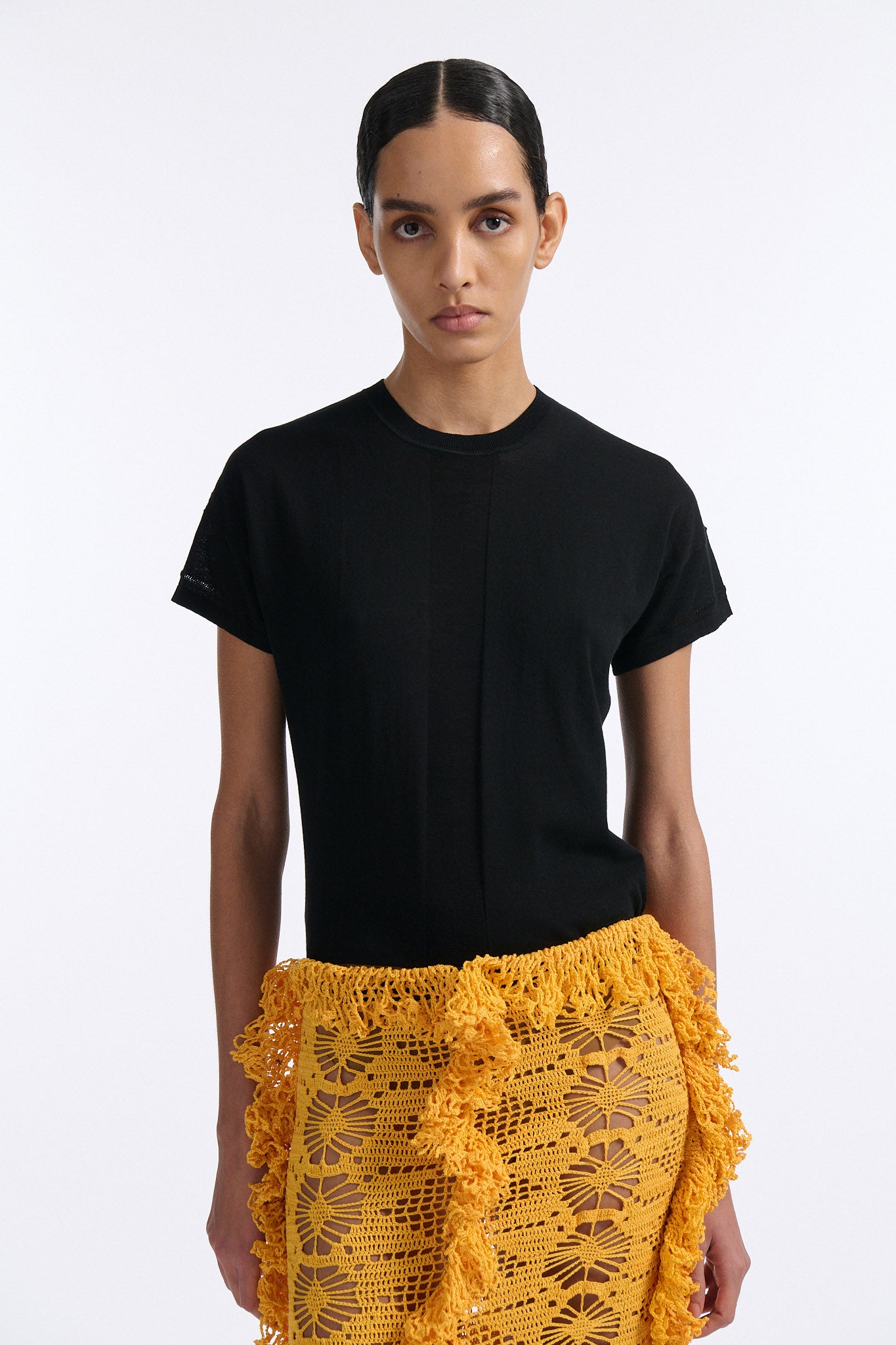 Female model with slicked back black hair in a bun wears black fine gauge cotton shirt with golden-yellow 4 panel crochet shirt. Skirt has elastic waist and crochet fringe finishing at all hems.