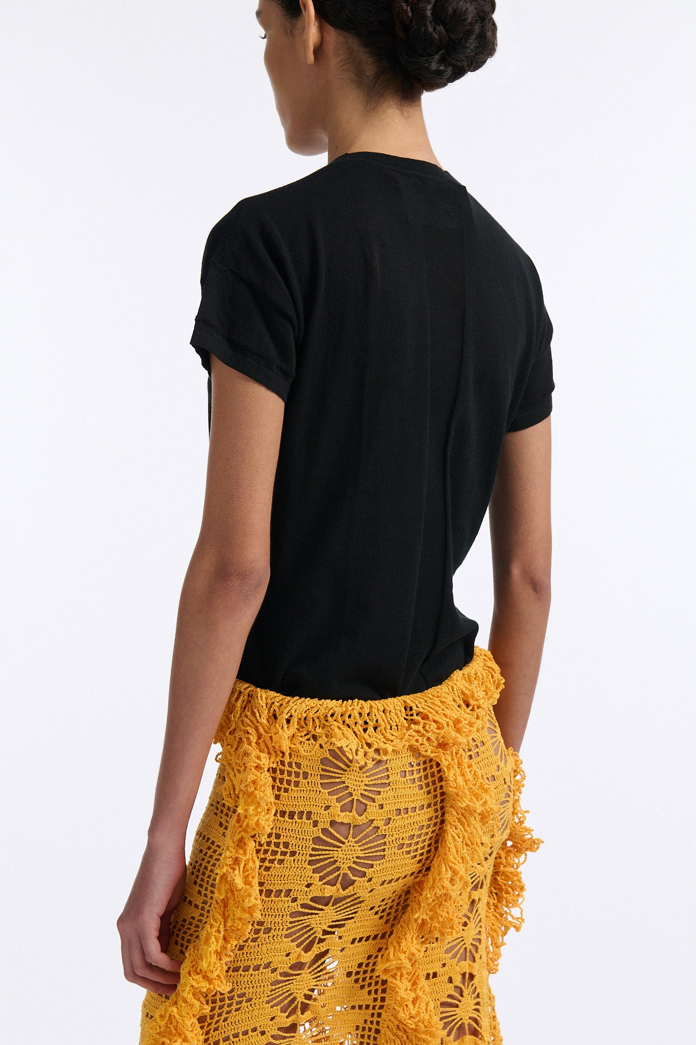 Female model with slicked back black hair in a bun wears black fine gauge cotton shirt with golden-yellow 4 panel crochet shirt. Skirt has elastic waist and crochet fringe finishing at all hems.