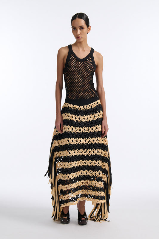 Female model with slicked back black hair in a bun wears black and ivory horizontal striped macrame skirt with asymmetric fringe on both sides. Model also wears fine mesh black crystal tank top.