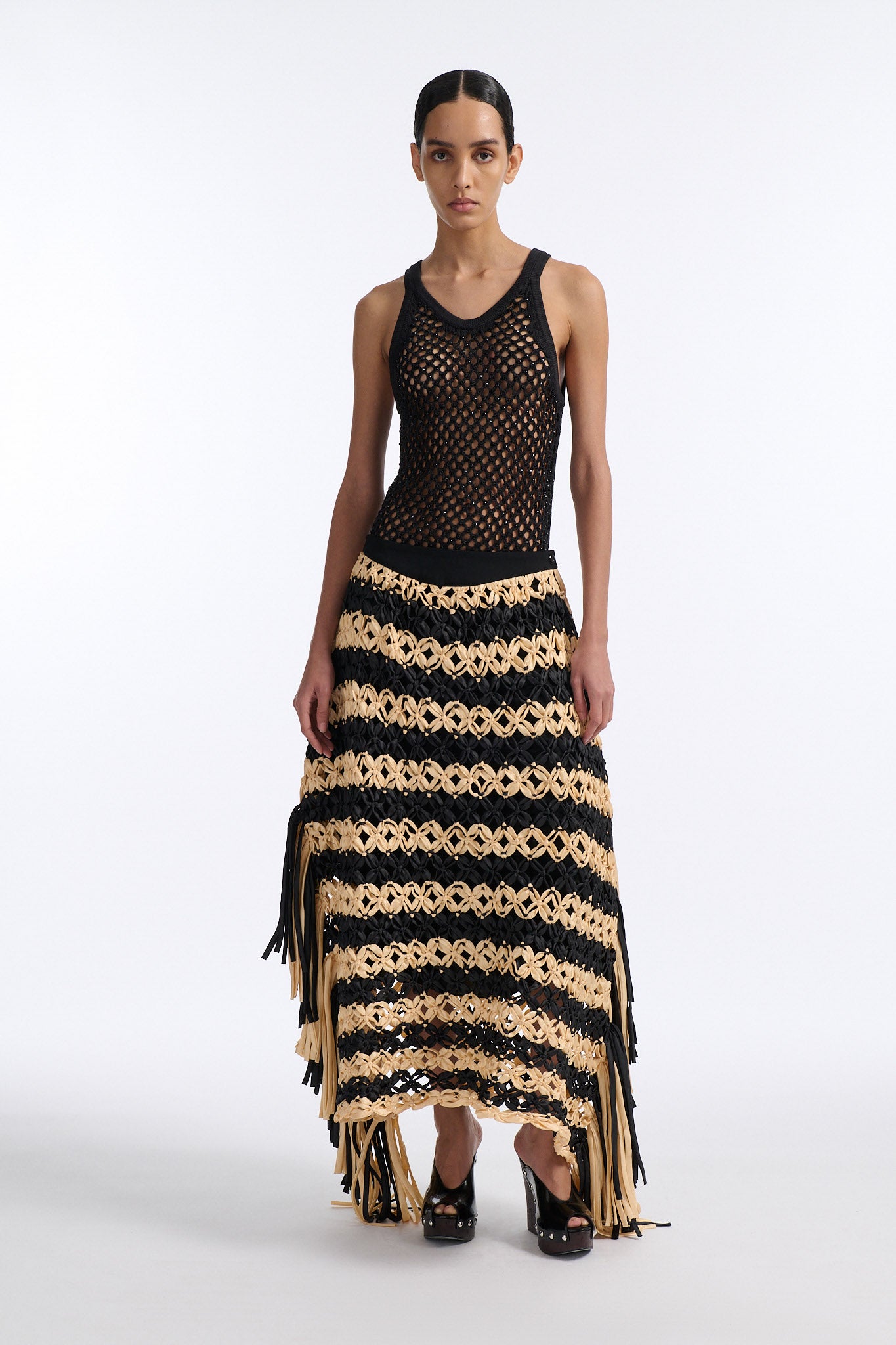 Female model with slicked back black hair in a bun wears black and ivory horizontal striped macrame skirt with asymmetric fringe on both sides. Model also wears fine mesh black crystal tank top.
