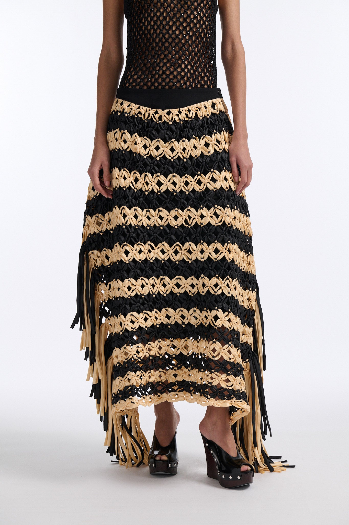 Female model with slicked back black hair in a bun wears black and ivory horizontal striped macrame skirt with asymmetric fringe on both sides. Model also wears fine mesh black crystal tank top.