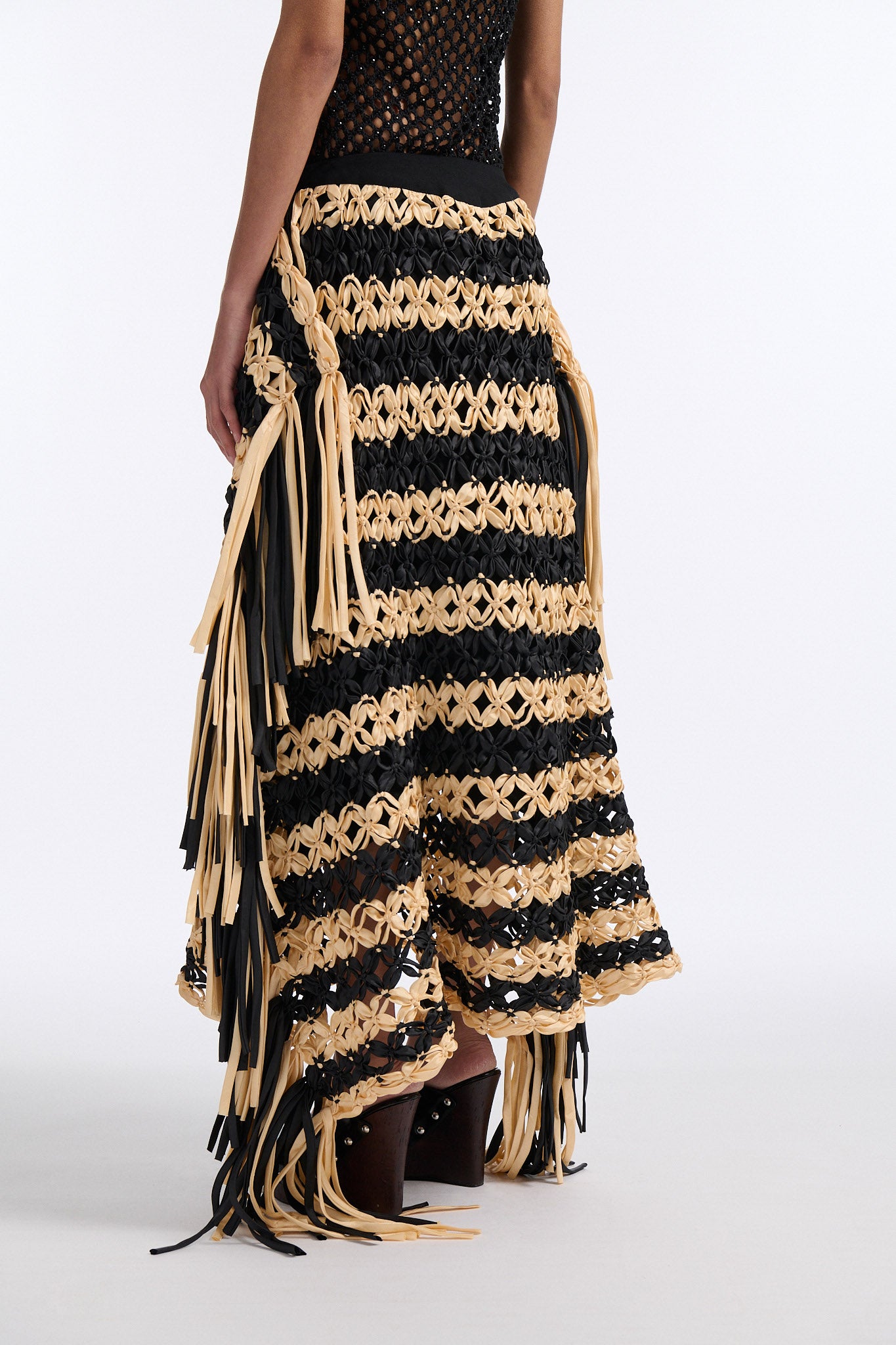 Female model with slicked back black hair in a bun wears black and ivory horizontal striped macrame skirt with asymmetric fringe on both sides. Model also wears fine mesh black crystal tank top.
