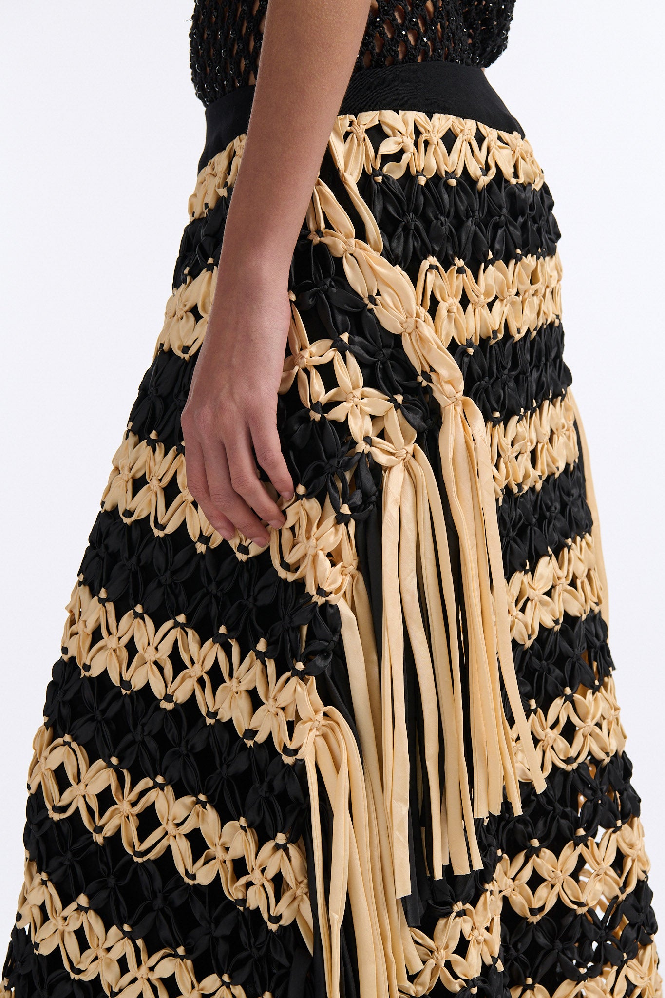 Female model with slicked back black hair in a bun wears black and ivory horizontal striped macrame skirt with asymmetric fringe on both sides. Model also wears fine mesh black crystal tank top.