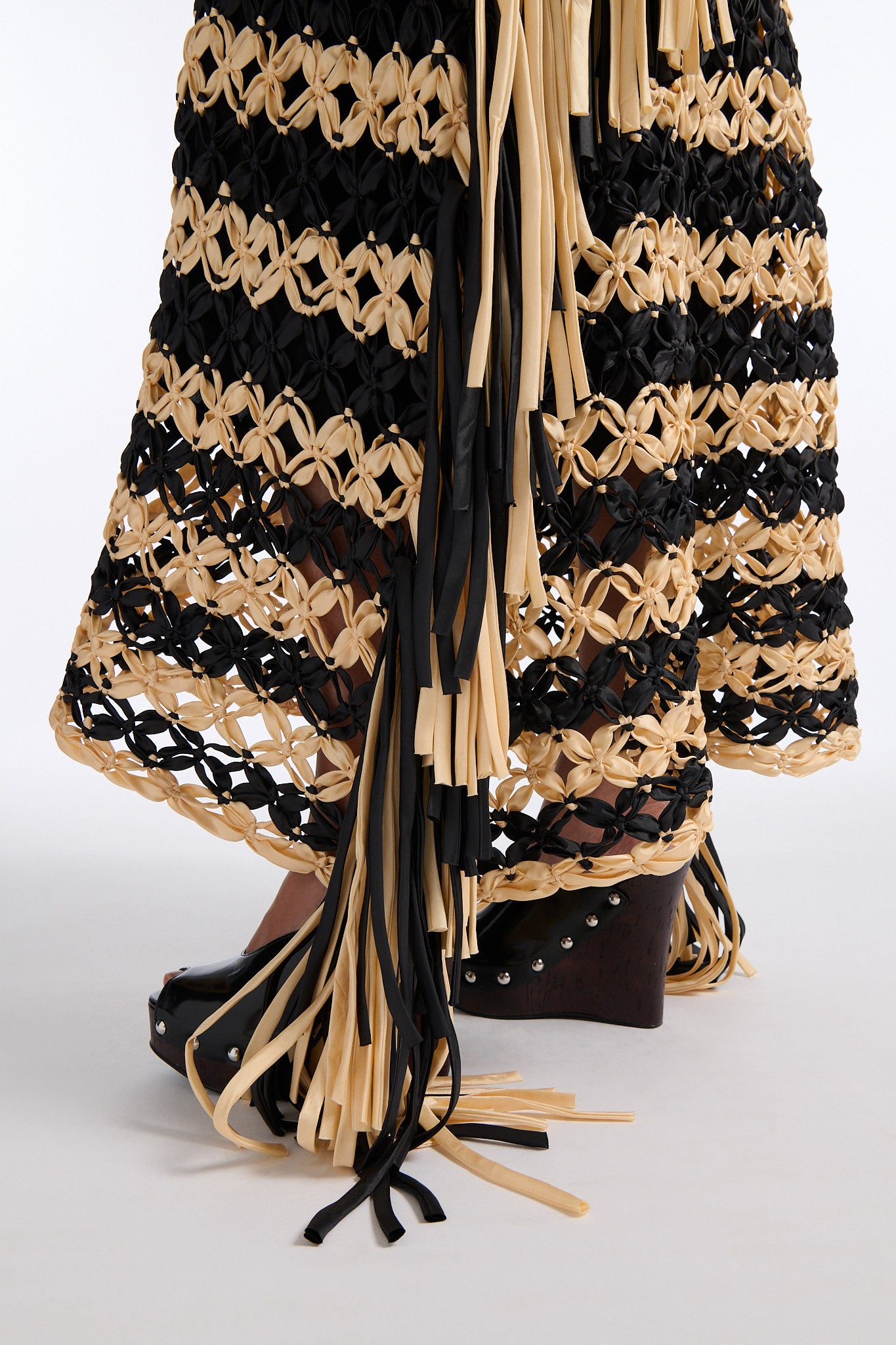 Female model with slicked back black hair in a bun wears black and ivory horizontal striped macrame skirt with asymmetric fringe on both sides. Model also wears fine mesh black crystal tank top.