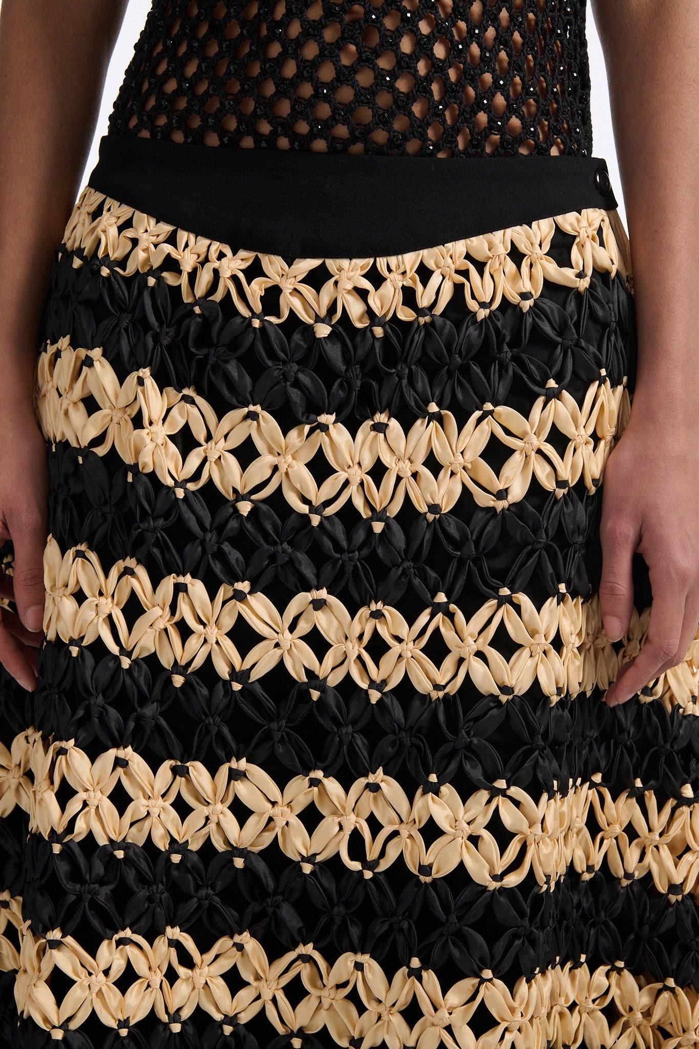 Female model with slicked back black hair in a bun wears black and ivory horizontal striped macrame skirt with asymmetric fringe on both sides. Model also wears fine mesh black crystal tank top.