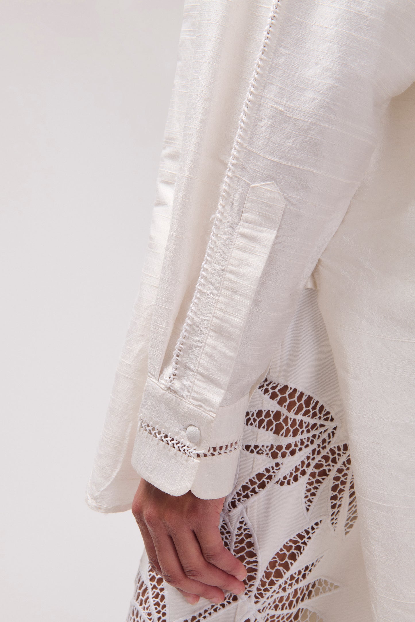 Close-up of a white textured silk shirt with a sleeve, button cuff, andpants with floral lace embroidery, shown on a model against a light neutral background.