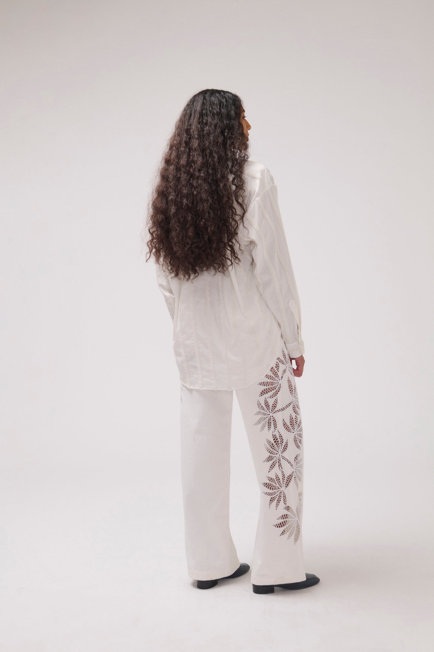 A woman with long, curly dark hair is shown from behind wearing a white long-sleeve shirt and matching wide-leg pants with brown leaf embroidery down the side of the pants, set against a plain light gray studio background.