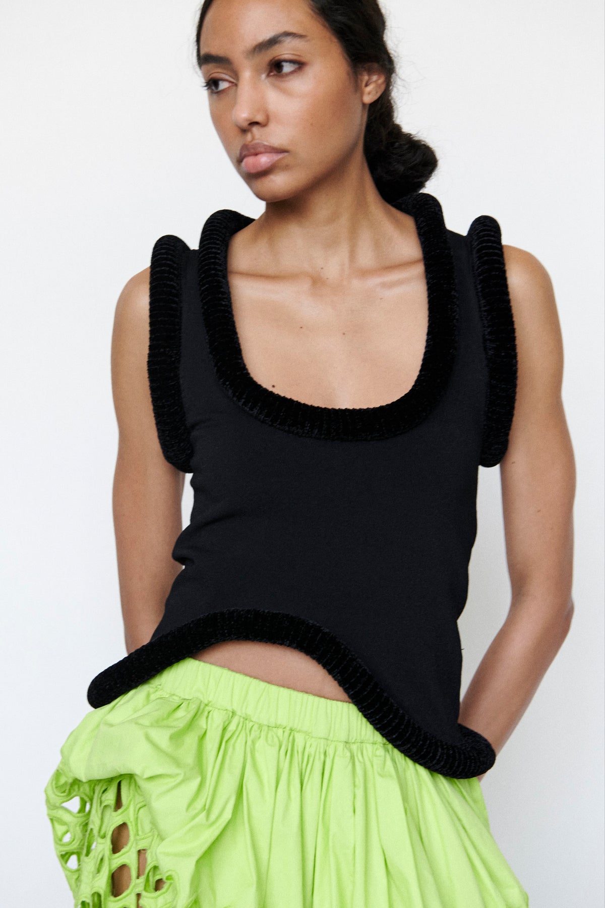 Model wearing black tank top with a plunge neckline and black sculptural piping around the neckline, sleeves and hem.