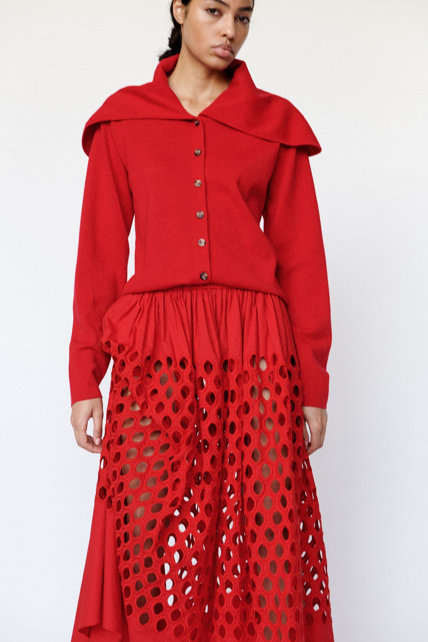 Model wearing long-sleeve red flat-knit button-down cardigan with exaggerated sailor collar. Model also wears perforated broderie anglaise gathered skirt