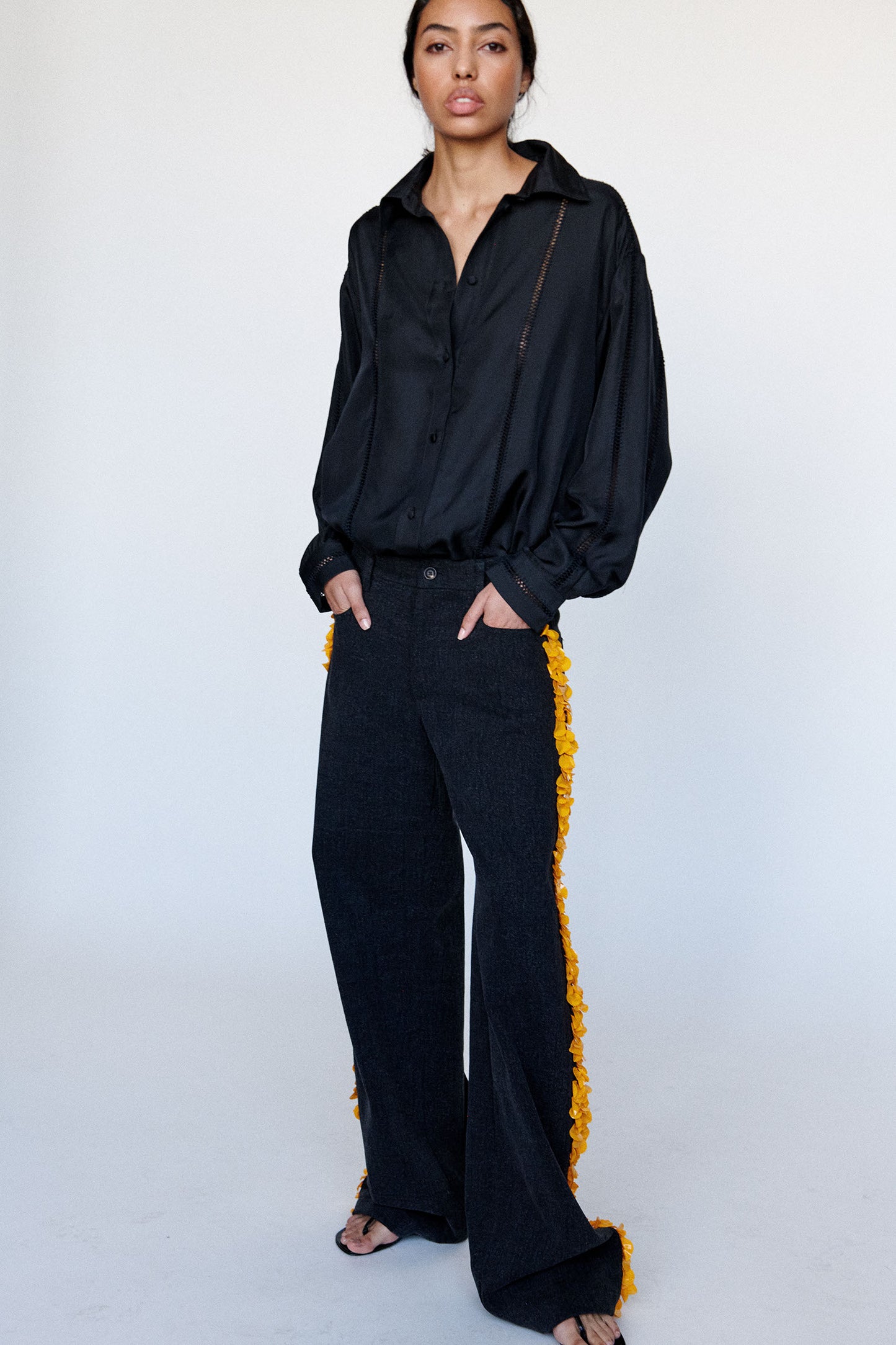 Model wears black silk shirt and black denim trousers with yellow paillette embellishments on both legs.