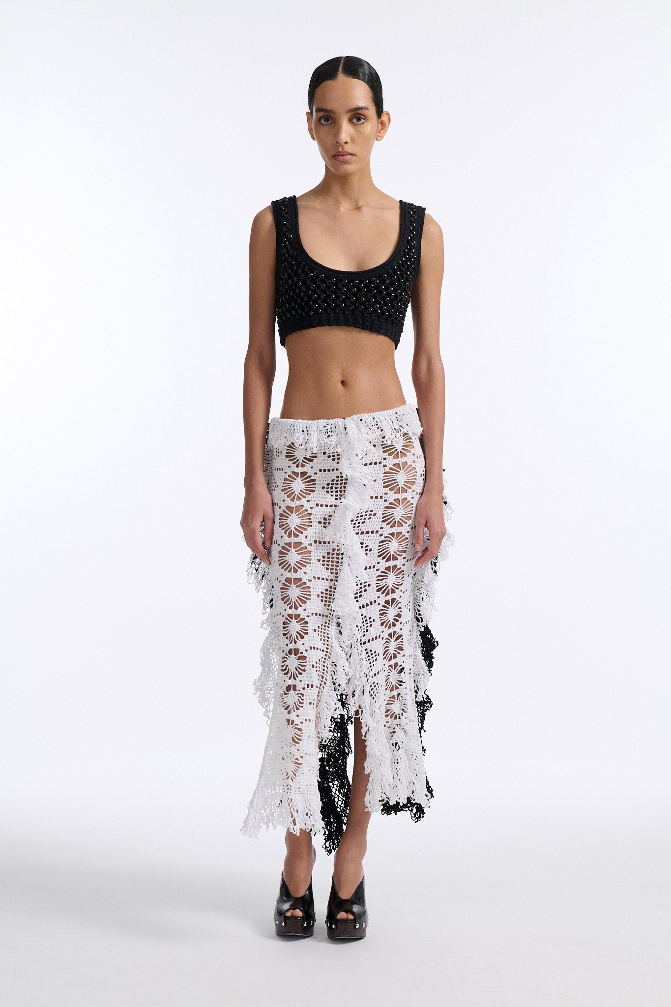 Female model with slicked back black hair in a bun wears black mesh bralette with four panel long crochet skirt. Crochet skirt has two white front panels and two black back panels.