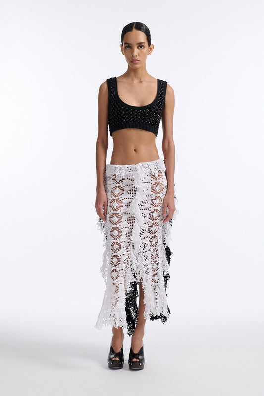 Female model with slicked back black hair in a bun wears black mesh bralette with four panel long crochet skirt. Crochet skirt has two white front panels and two black back panels.