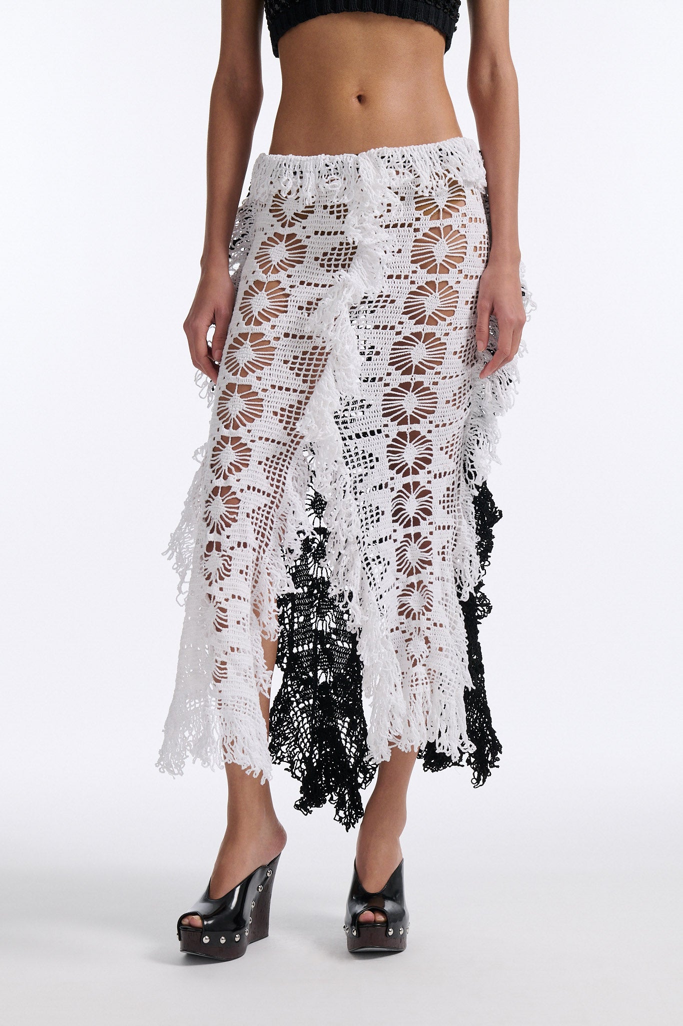 Female model with slicked back black hair in a bun wears black mesh bralette with four panel long crochet skirt. Crochet skirt has two white front panels and two black back panels.