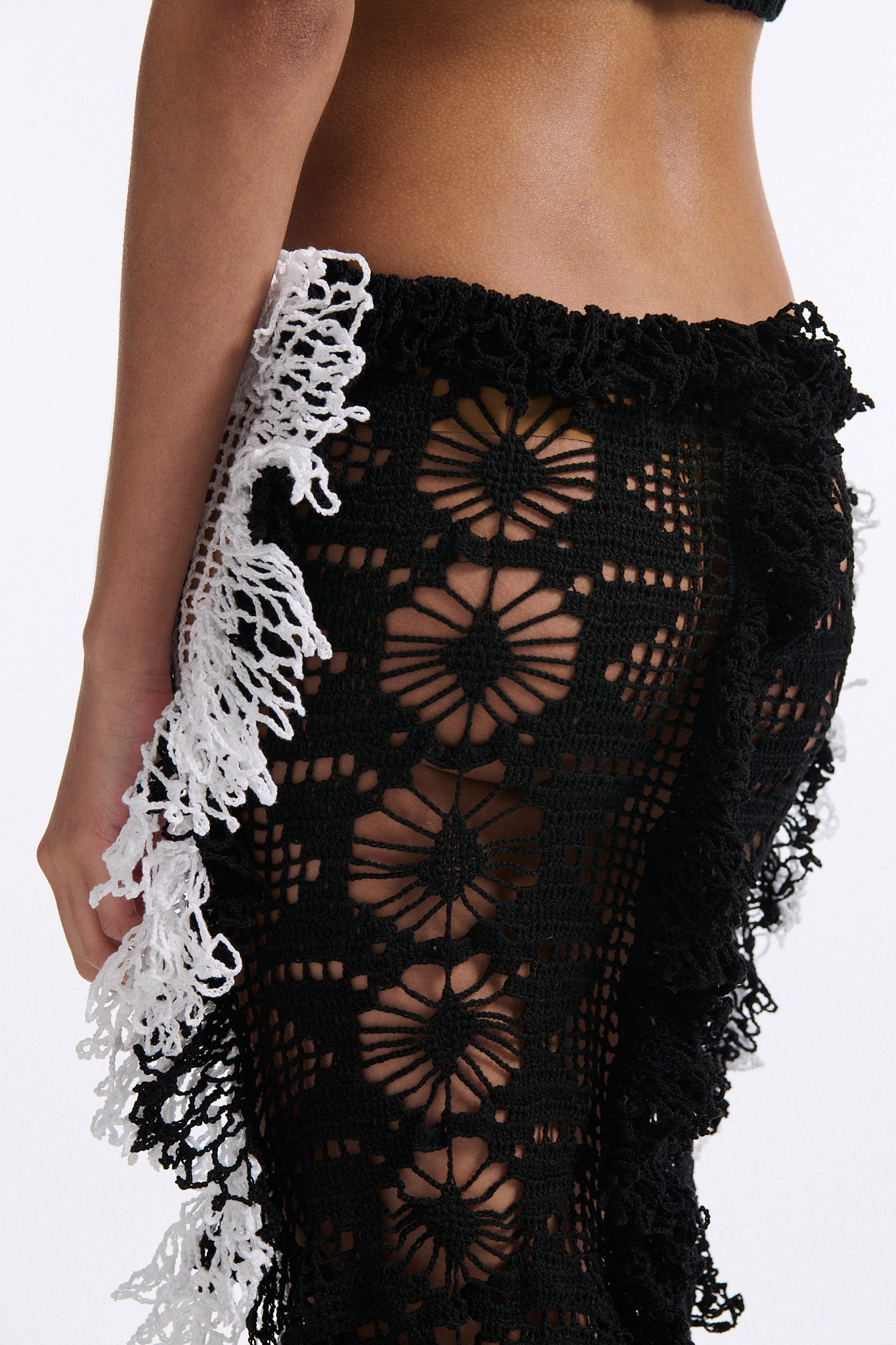 Female model with slicked back black hair in a bun wears black mesh bralette with four panel long crochet skirt. Crochet skirt has two white front panels and two black back panels.