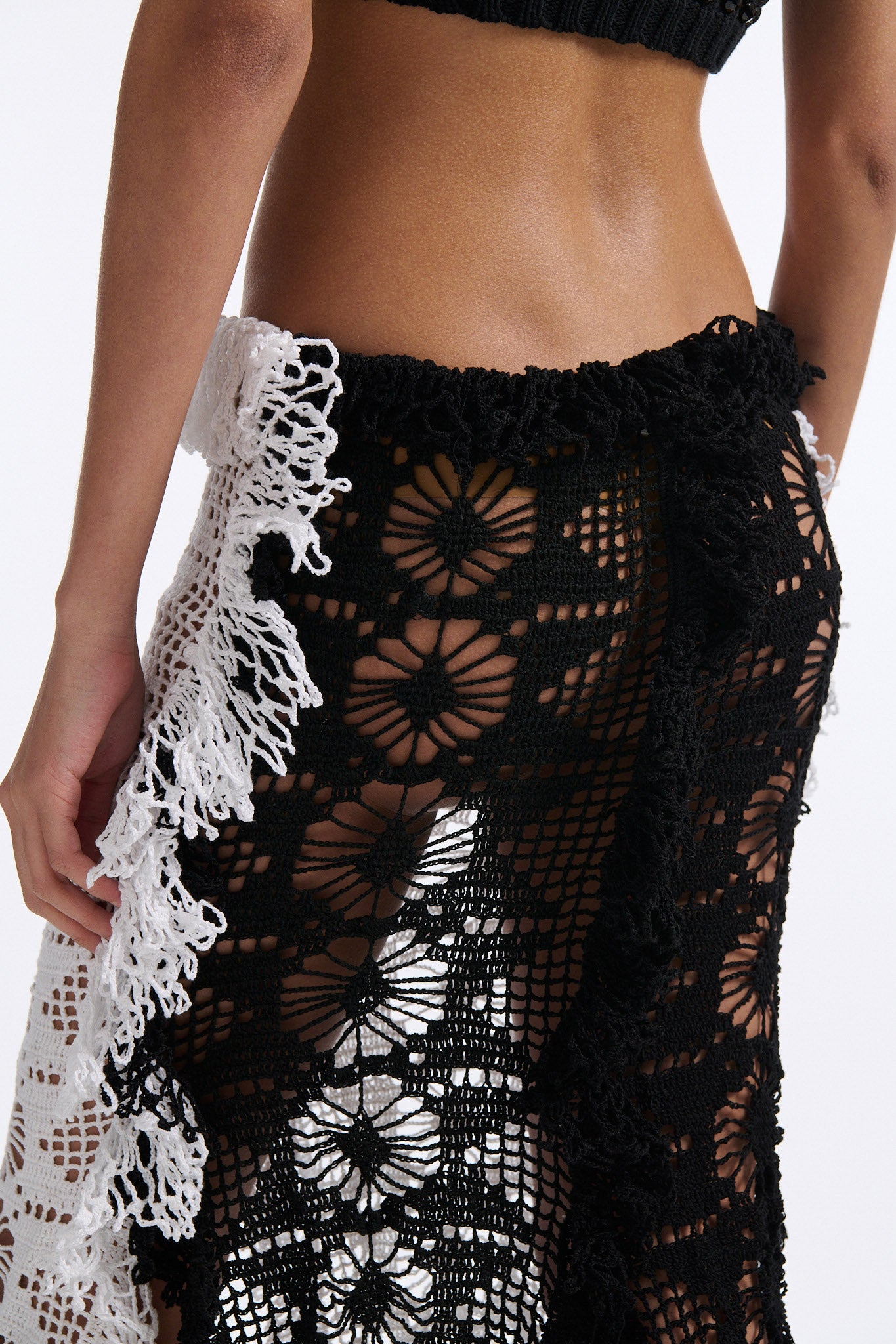 Female model with slicked back black hair in a bun wears black mesh bralette with four panel long crochet skirt. Crochet skirt has two white front panels and two black back panels.
