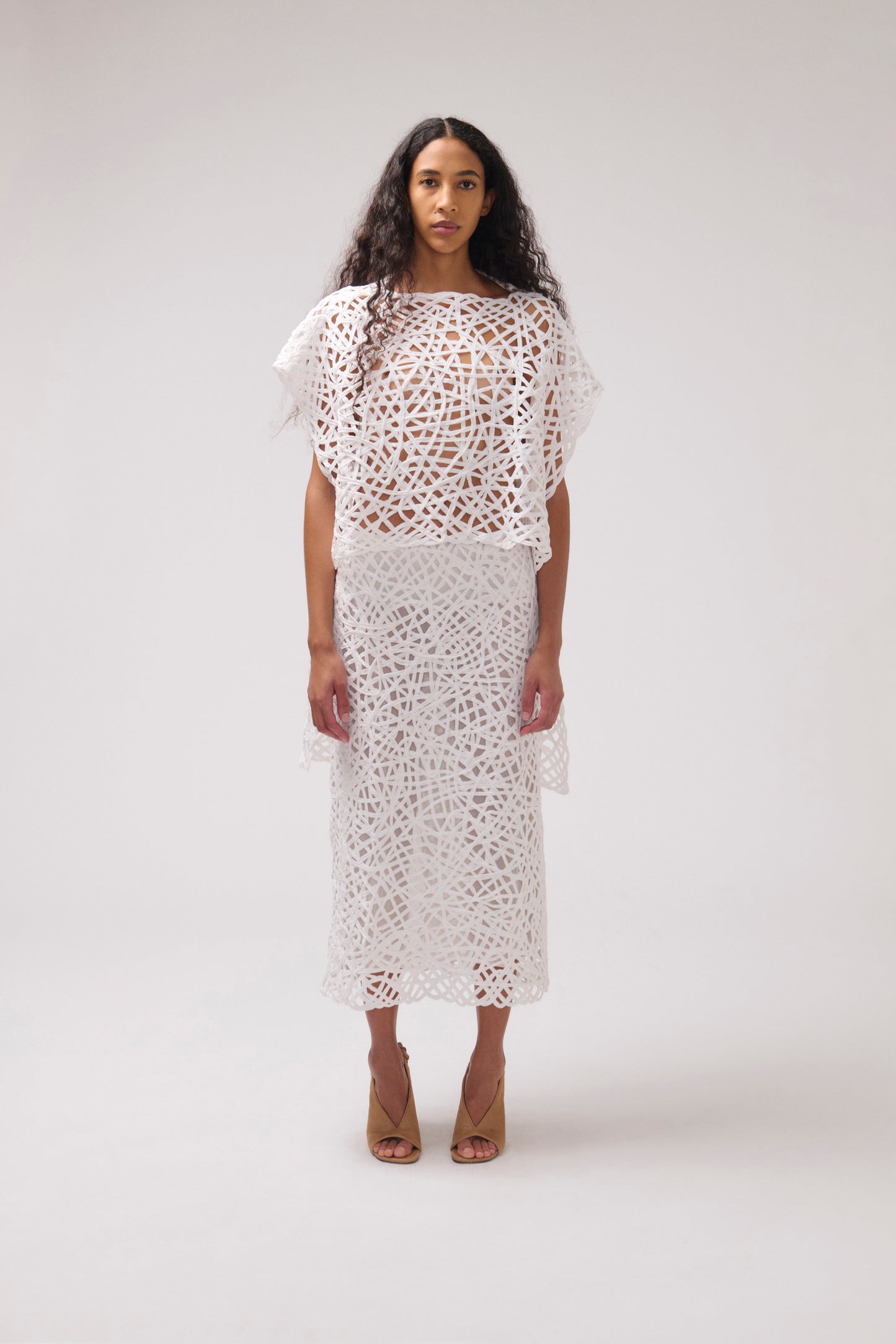 A model stands against a light gray studio background wearing a white crochet or lace two-piece outfit with a short flutter-sleeve top and a midi skirt, paired with tan heeled sandals.