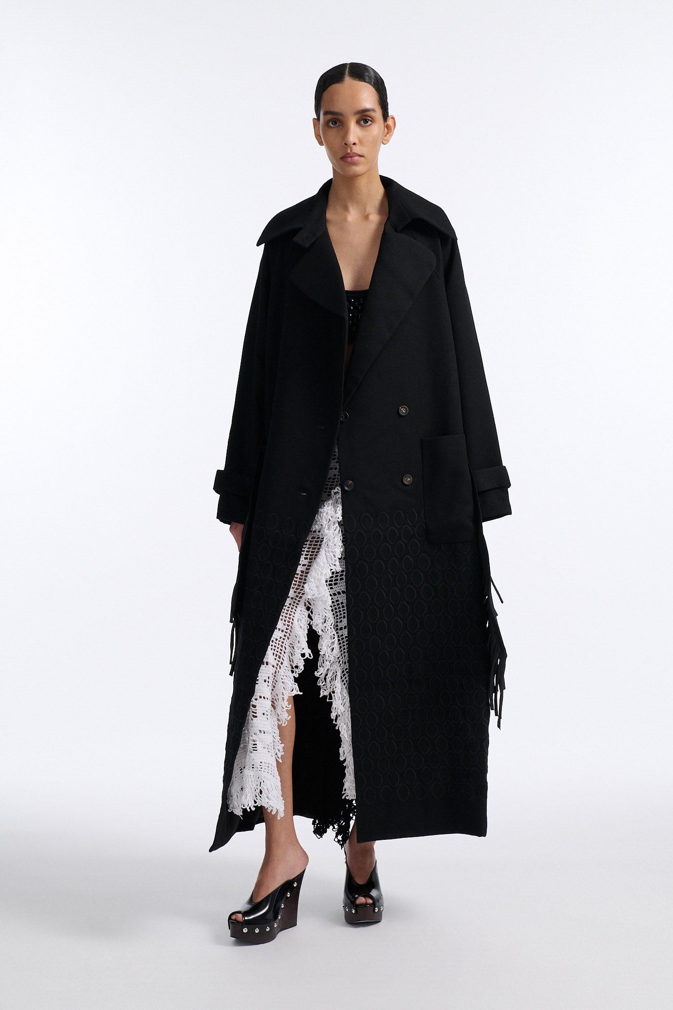 Female model with slicked back black hair in a bun wears long black wool coat with oversized collar and fringe belt.