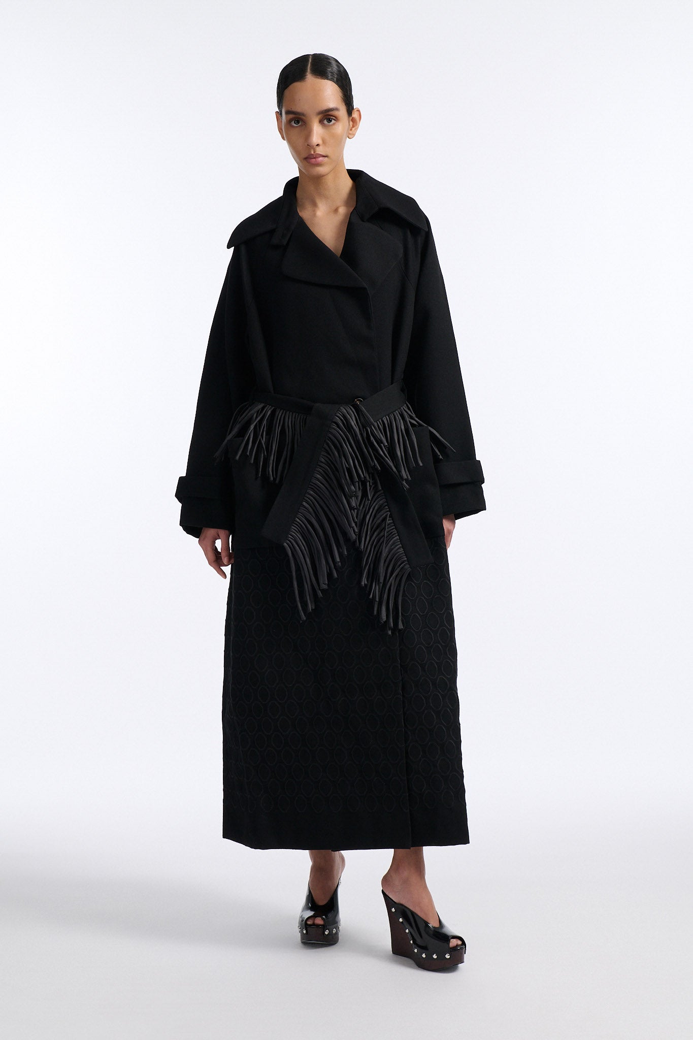 Female model with slicked back black hair in a bun wears long black wool coat with oversized collar and fringe belt.