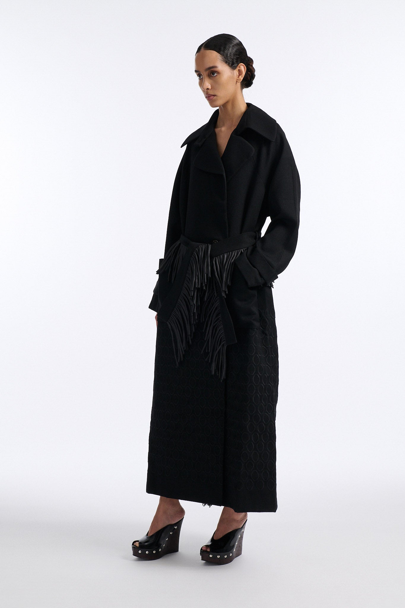Female model with slicked back black hair in a bun wears long black wool coat with oversized collar and fringe belt.