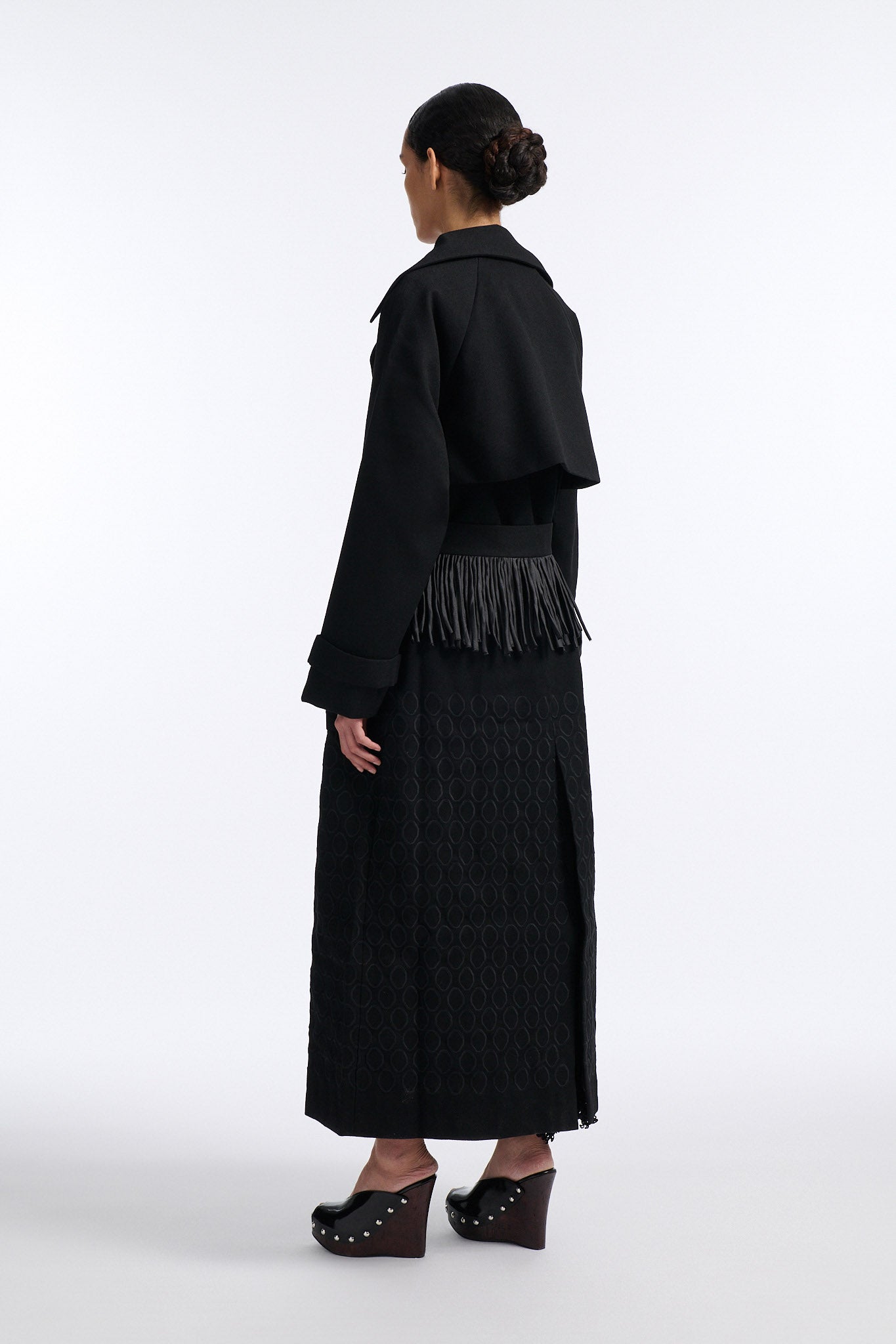 Female model with slicked back black hair in a bun wears long black wool coat with oversized collar and fringe belt.