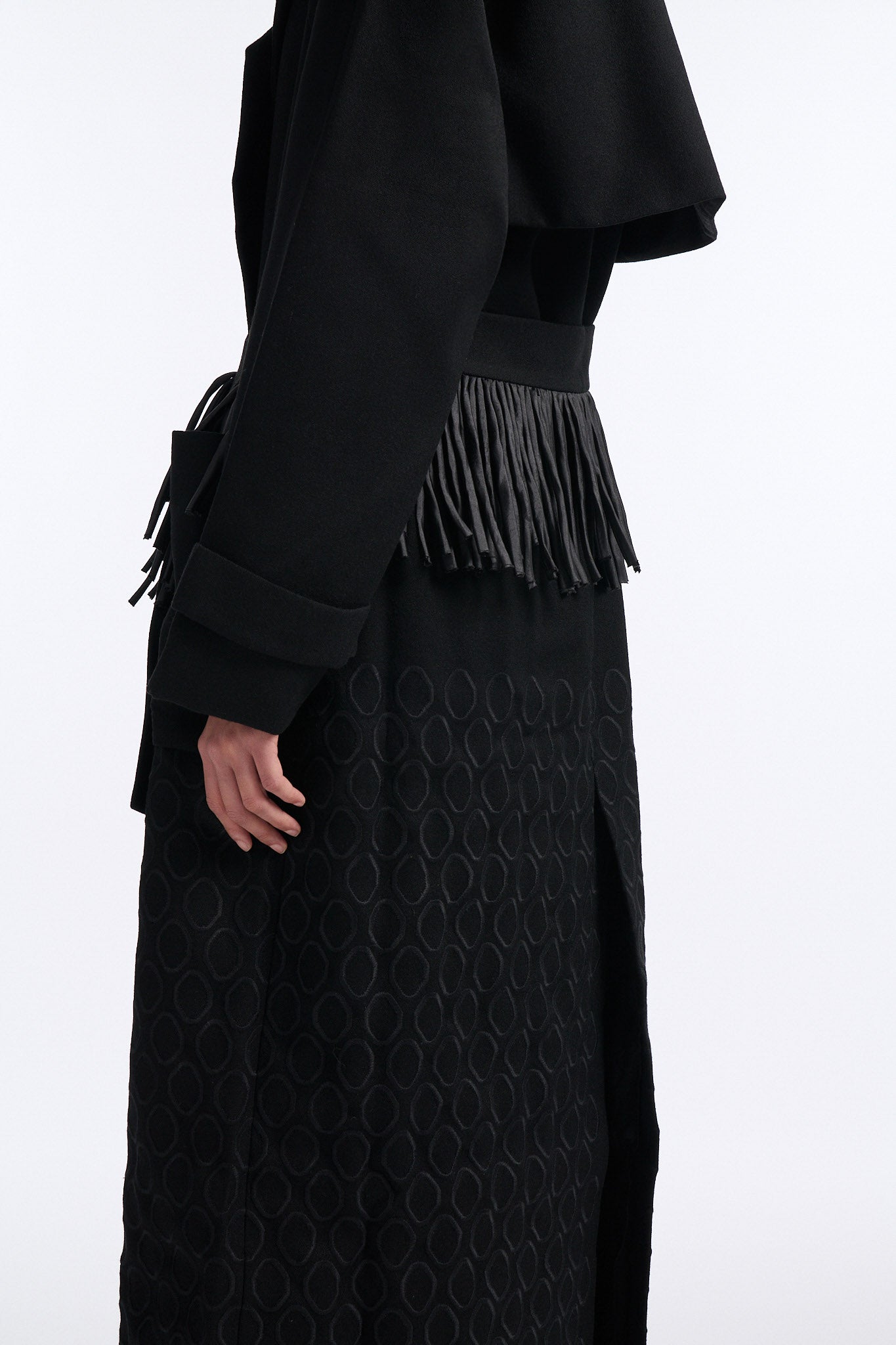 Female model with slicked back black hair in a bun wears long black wool coat with oversized collar and fringe belt.