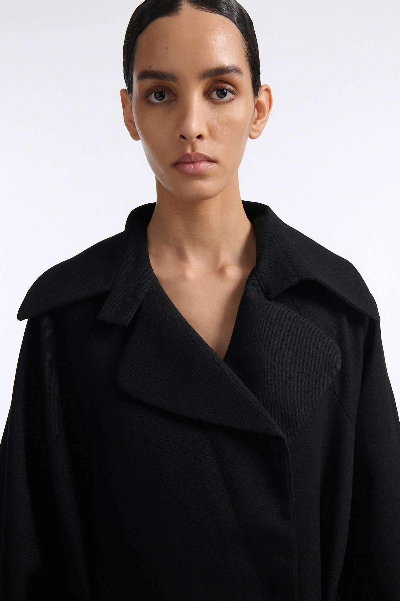 Female model with slicked back black hair in a bun wears long black wool coat with oversized collar and fringe belt.