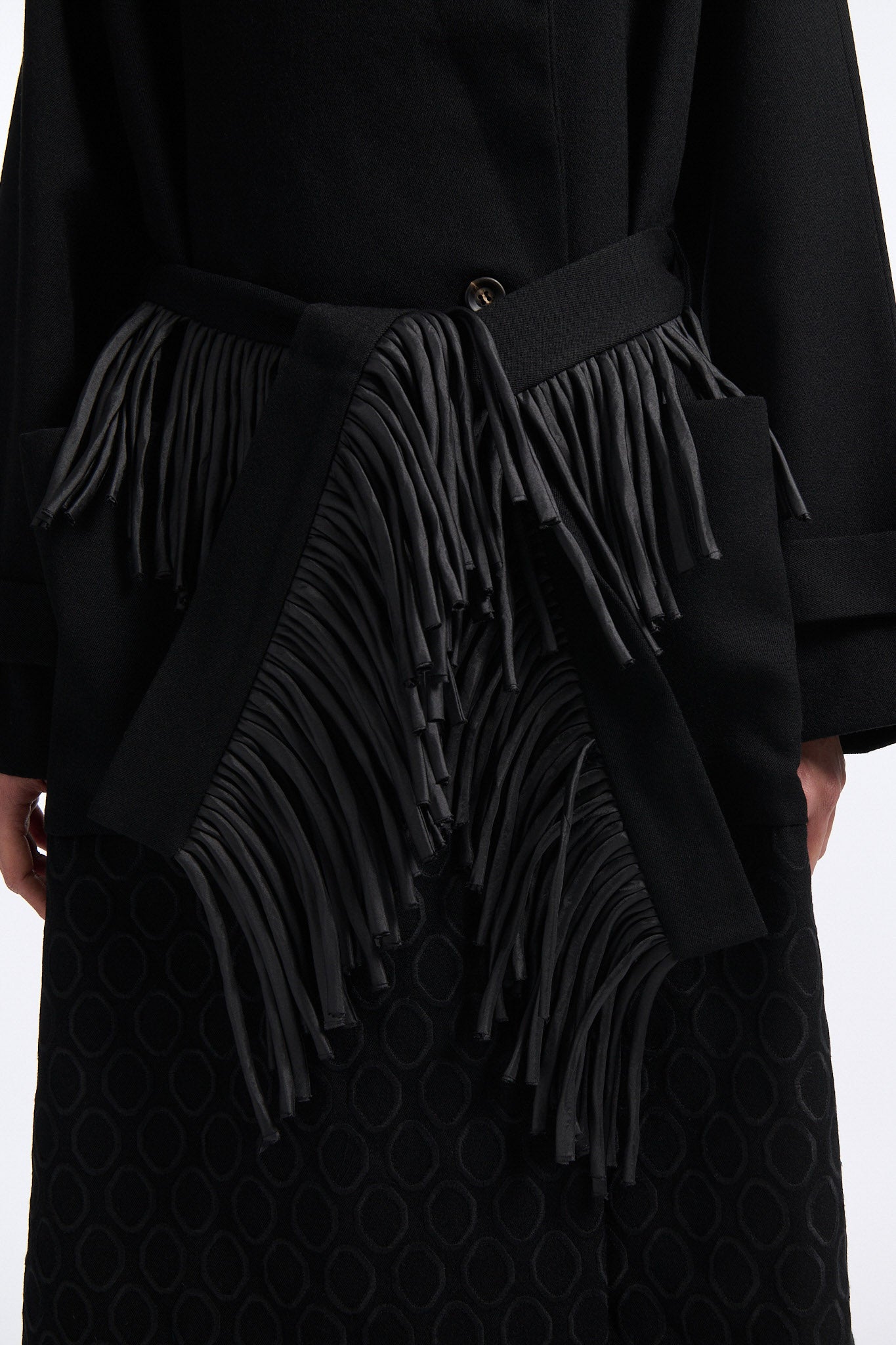 Female model with slicked back black hair in a bun wears long black wool coat with oversized collar and fringe belt.