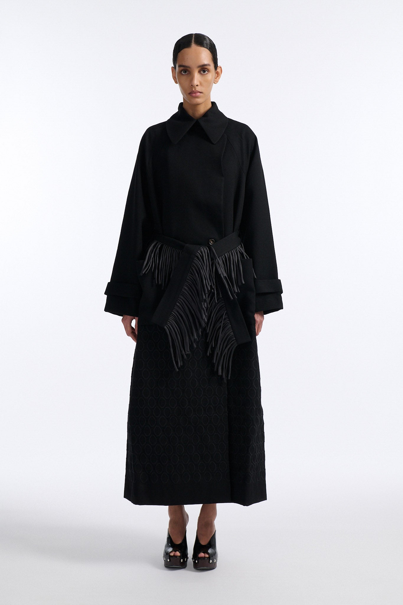 Female model with slicked back black hair in a bun wears long black wool coat with oversized collar and fringe belt.
