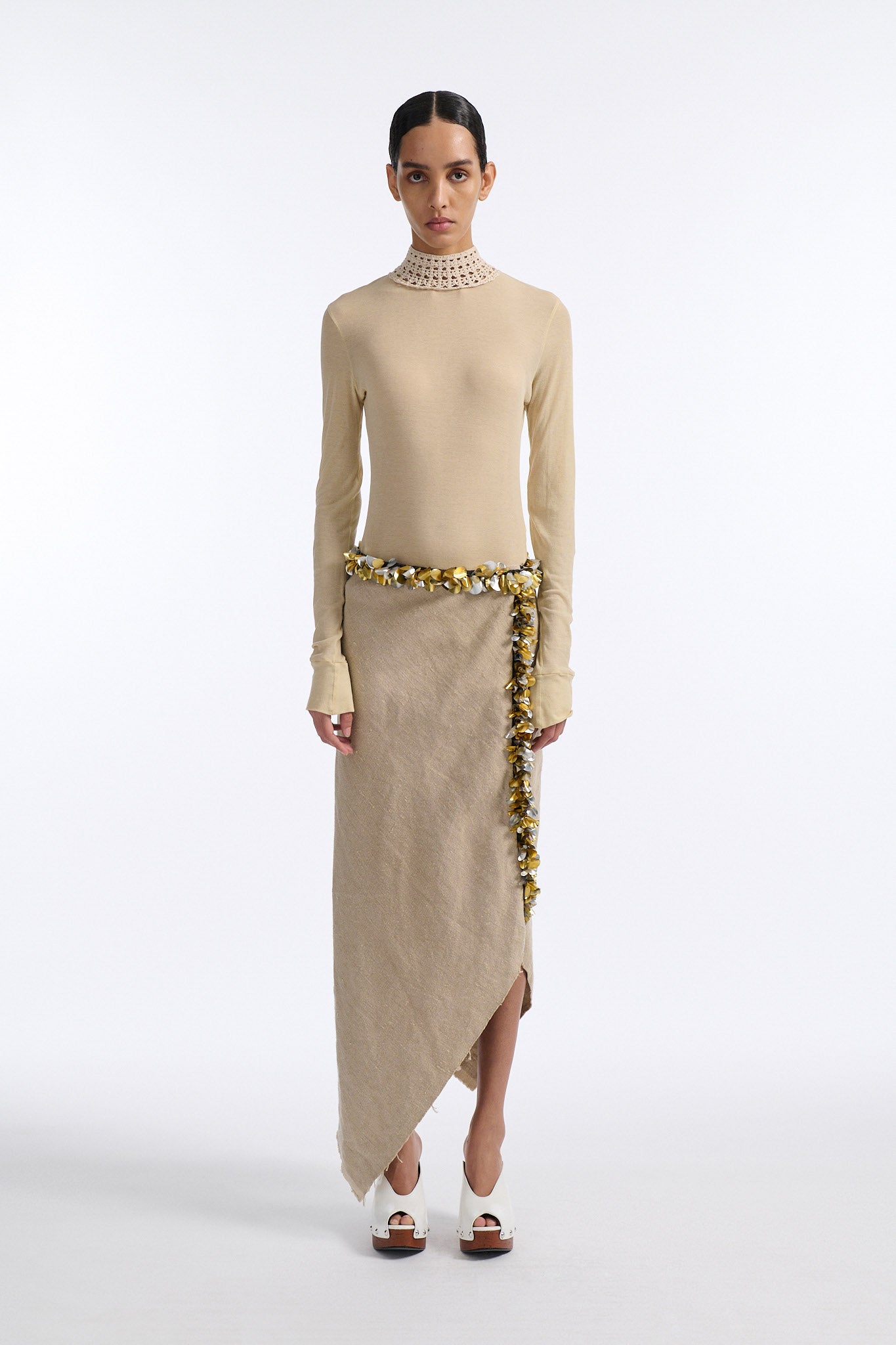 Female model with slicked back black hair in a bun wears beige linen handkerchief wrap skirt. Waist and closure are lined with melted metallic embellishments in gold and silver to provide an oyster illusion. Model also wears light beige fine-gauge cotton long sleeve top with mockneck crochet collar.