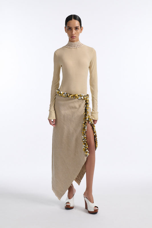 Female model with slicked back black hair in a bun wears beige linen handkerchief wrap skirt. Waist and closure are lined with melted metallic embellishments in gold and silver to provide an oyster illusion. Model also wears light beige fine-gauge cotton long sleeve top with mockneck crochet collar.