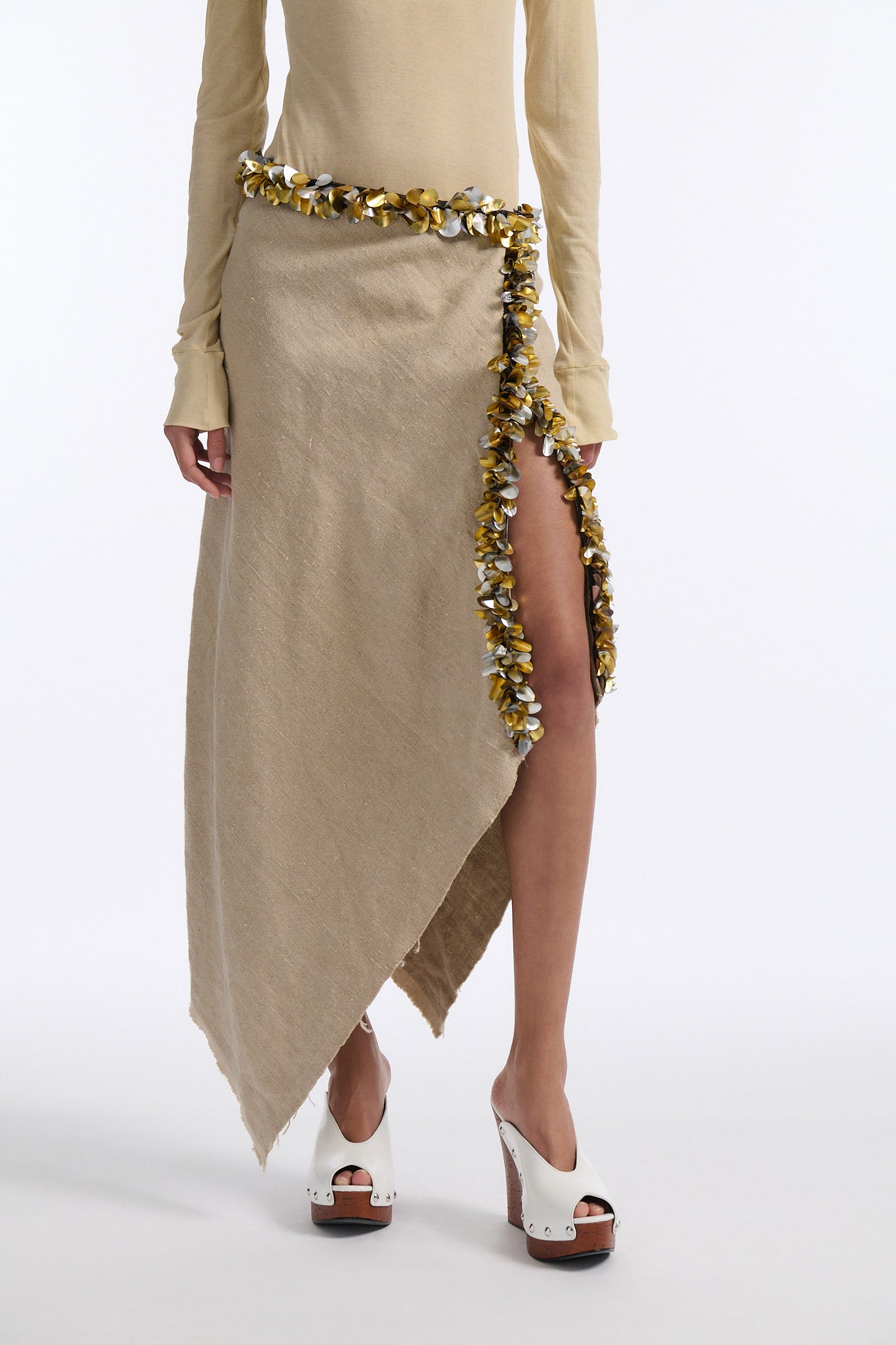Female model with slicked back black hair in a bun wears beige linen handkerchief wrap skirt. Waist and closure are lined with melted metallic embellishments in gold and silver to provide an oyster illusion. Model also wears light beige fine-gauge cotton long sleeve top with mockneck crochet collar.
