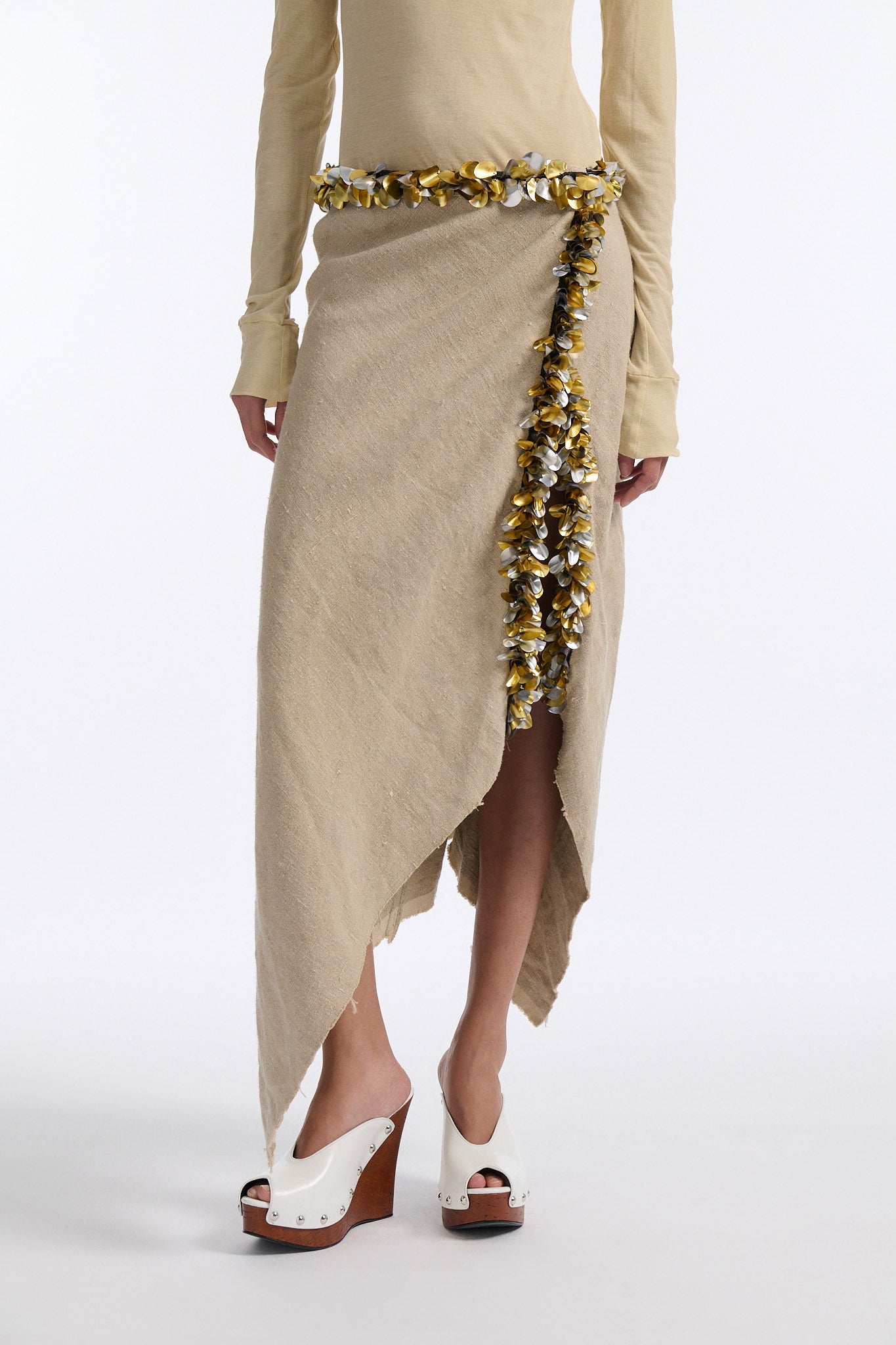 Female model with slicked back black hair in a bun wears beige linen handkerchief wrap skirt. Waist and closure are lined with melted metallic embellishments in gold and silver to provide an oyster illusion. Model also wears light beige fine-gauge cotton long sleeve top with mockneck crochet collar.