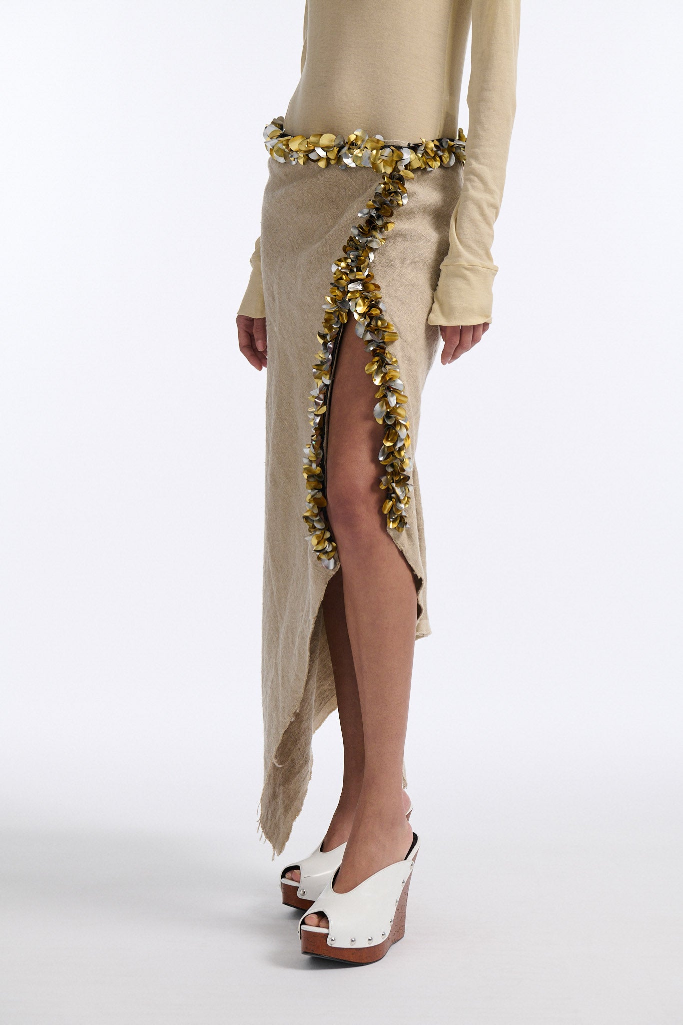 Female model with slicked back black hair in a bun wears beige linen handkerchief wrap skirt. Waist and closure are lined with melted metallic embellishments in gold and silver to provide an oyster illusion. Model also wears light beige fine-gauge cotton long sleeve top with mockneck crochet collar.