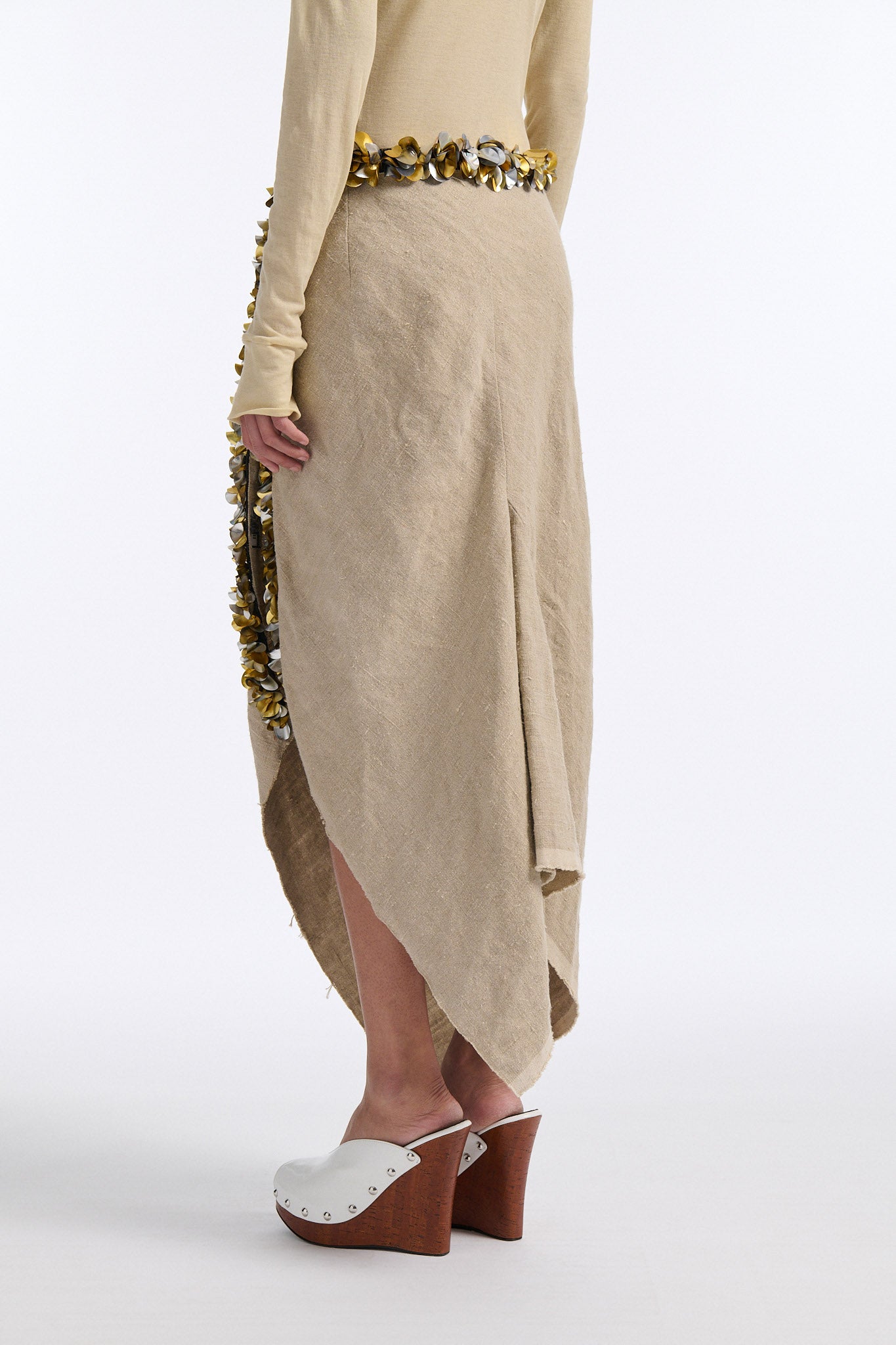 Female model with slicked back black hair in a bun wears beige linen handkerchief wrap skirt. Waist and closure are lined with melted metallic embellishments in gold and silver to provide an oyster illusion. Model also wears light beige fine-gauge cotton long sleeve top with mockneck crochet collar.