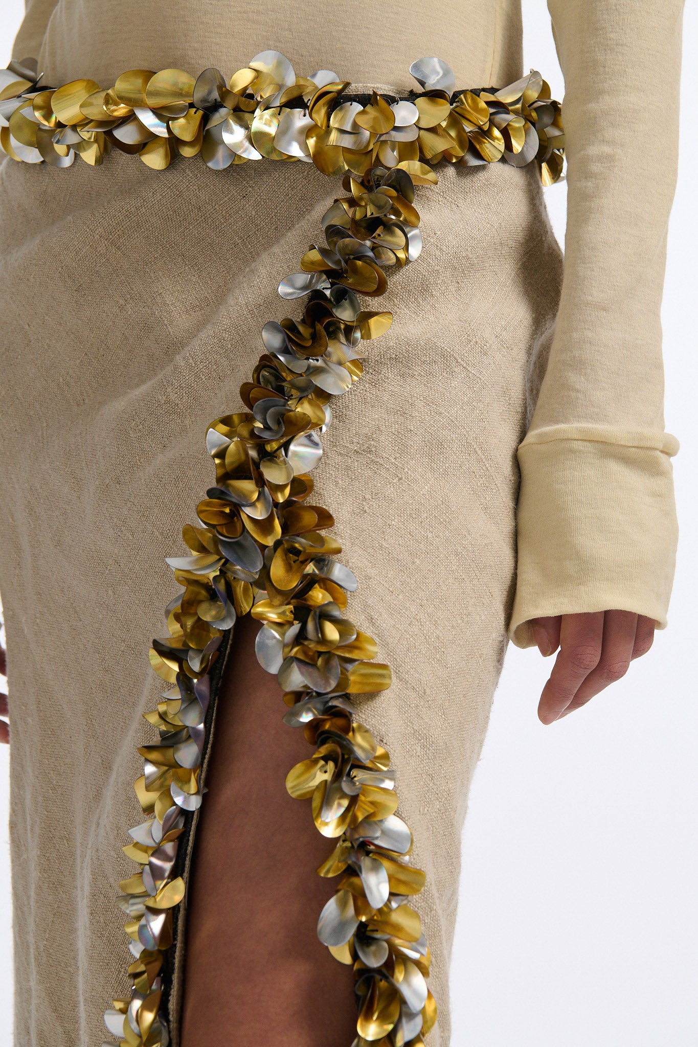 Female model with slicked back black hair in a bun wears beige linen handkerchief wrap skirt. Waist and closure are lined with melted metallic embellishments in gold and silver to provide an oyster illusion. Model also wears light beige fine-gauge cotton long sleeve top with mockneck crochet collar.