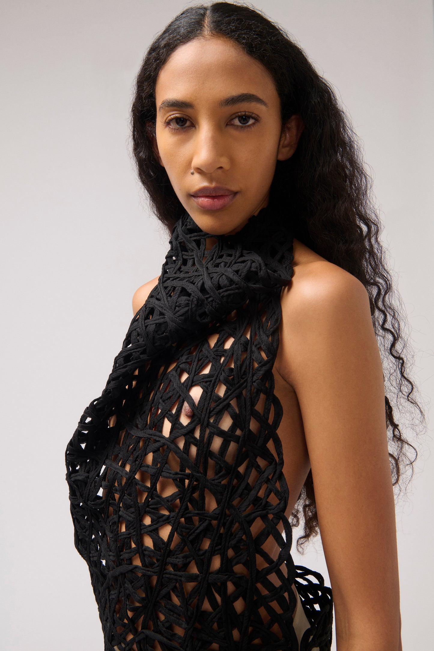 A woman with long, curly dark hair wearing a black open-knit crochet halter top and matching scarf against a plain light gray studio background.