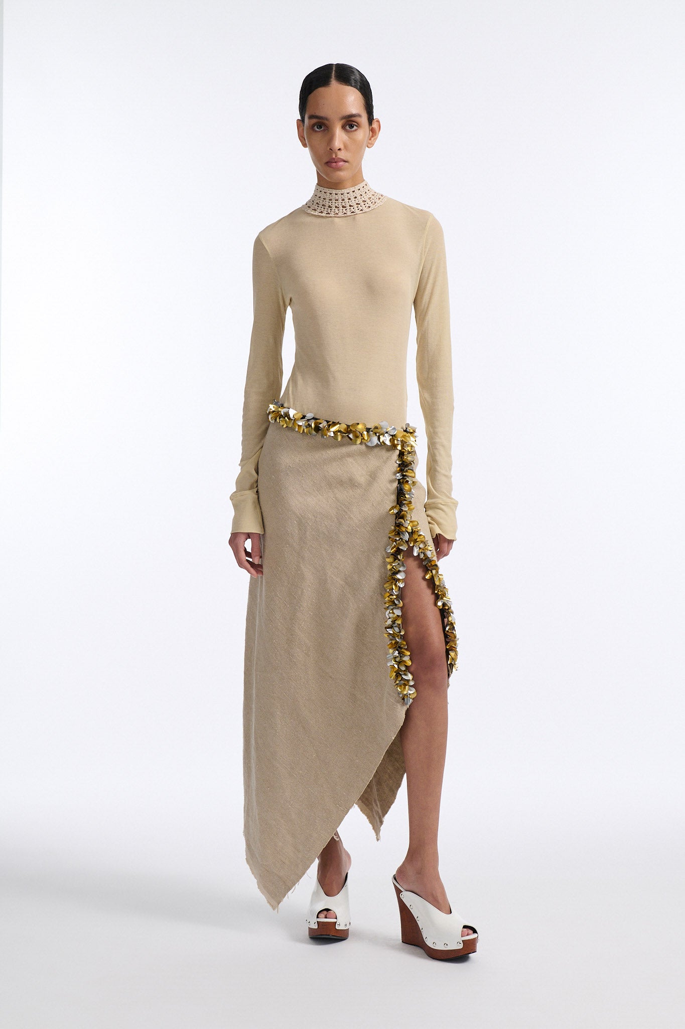 Female model with slicked back black hair in a bun wears beige linen handkerchief wrap skirt. Waist and closure are lined with melted metallic embellishments in gold and silver to provide an oyster illusion. Model also wears light beige fine-gauge cotton long sleeve top with mockneck crochet collar.