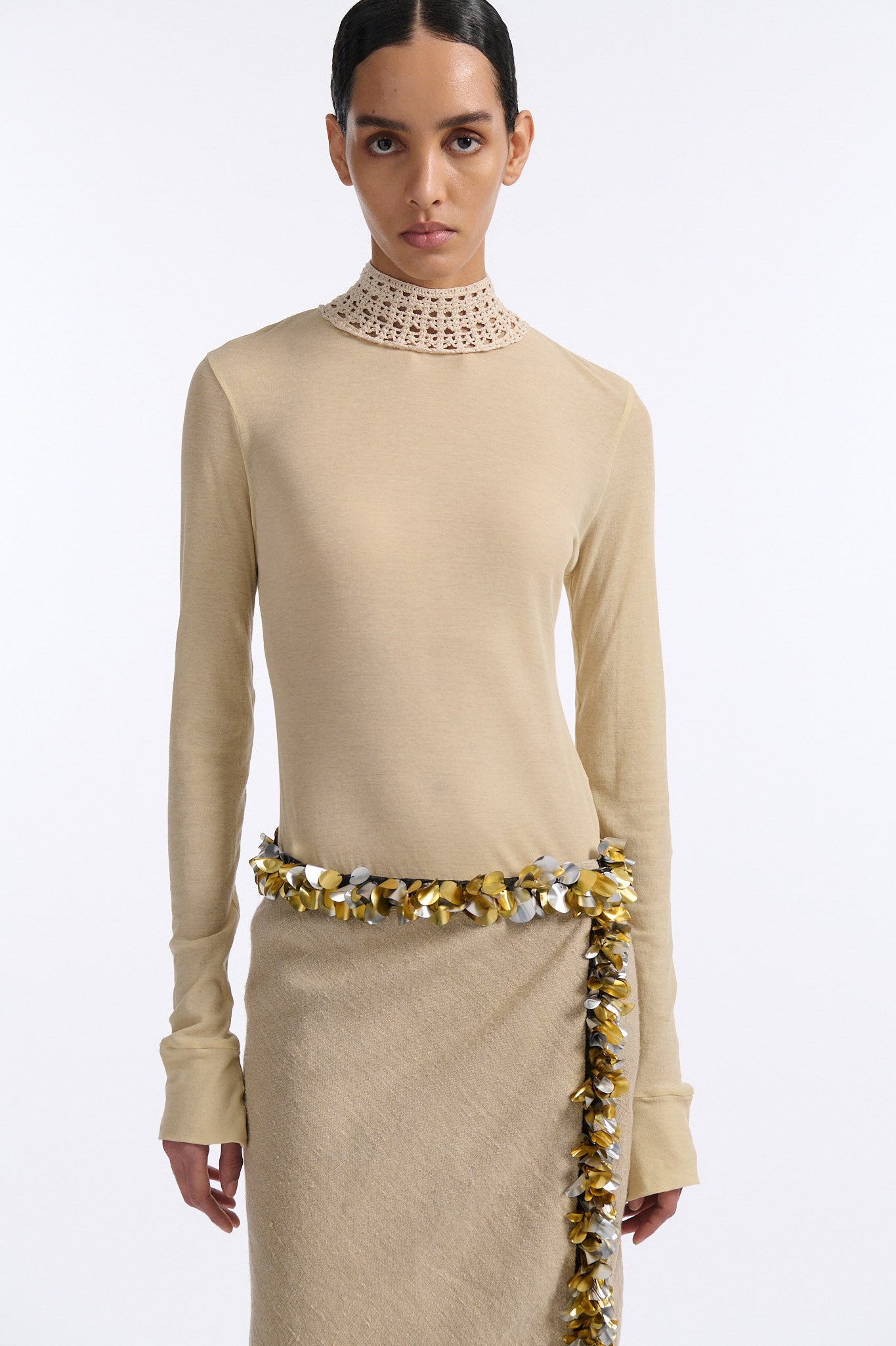 Female model with slicked back black hair in a bun wears beige linen handkerchief wrap skirt. Waist and closure are lined with melted metallic embellishments in gold and silver to provide an oyster illusion. Model also wears light beige fine-gauge cotton long sleeve top with mockneck crochet collar.