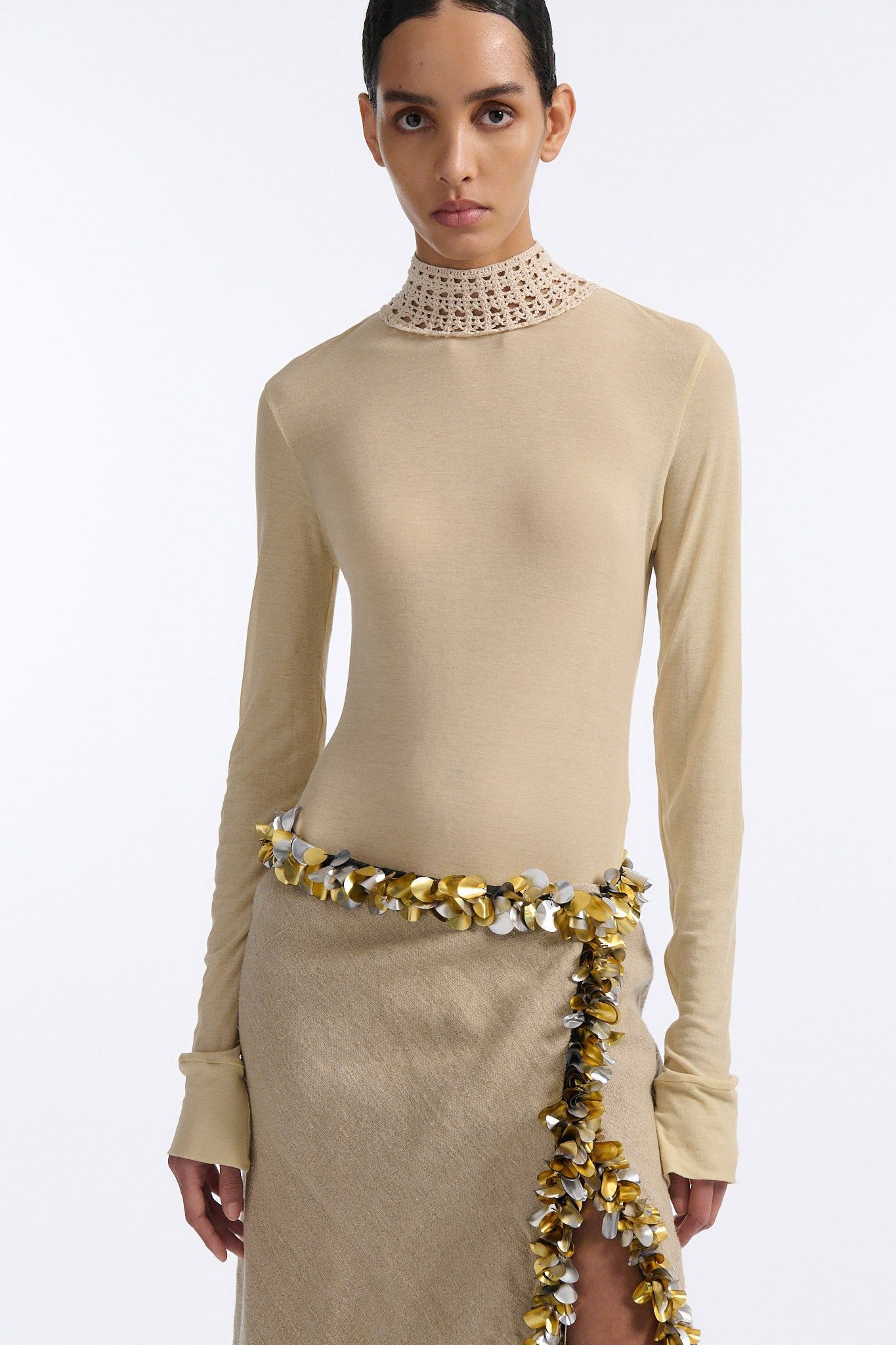 Female model with slicked back black hair in a bun wears beige linen handkerchief wrap skirt. Waist and closure are lined with melted metallic embellishments in gold and silver to provide an oyster illusion. Model also wears light beige fine-gauge cotton long sleeve top with mockneck crochet collar.