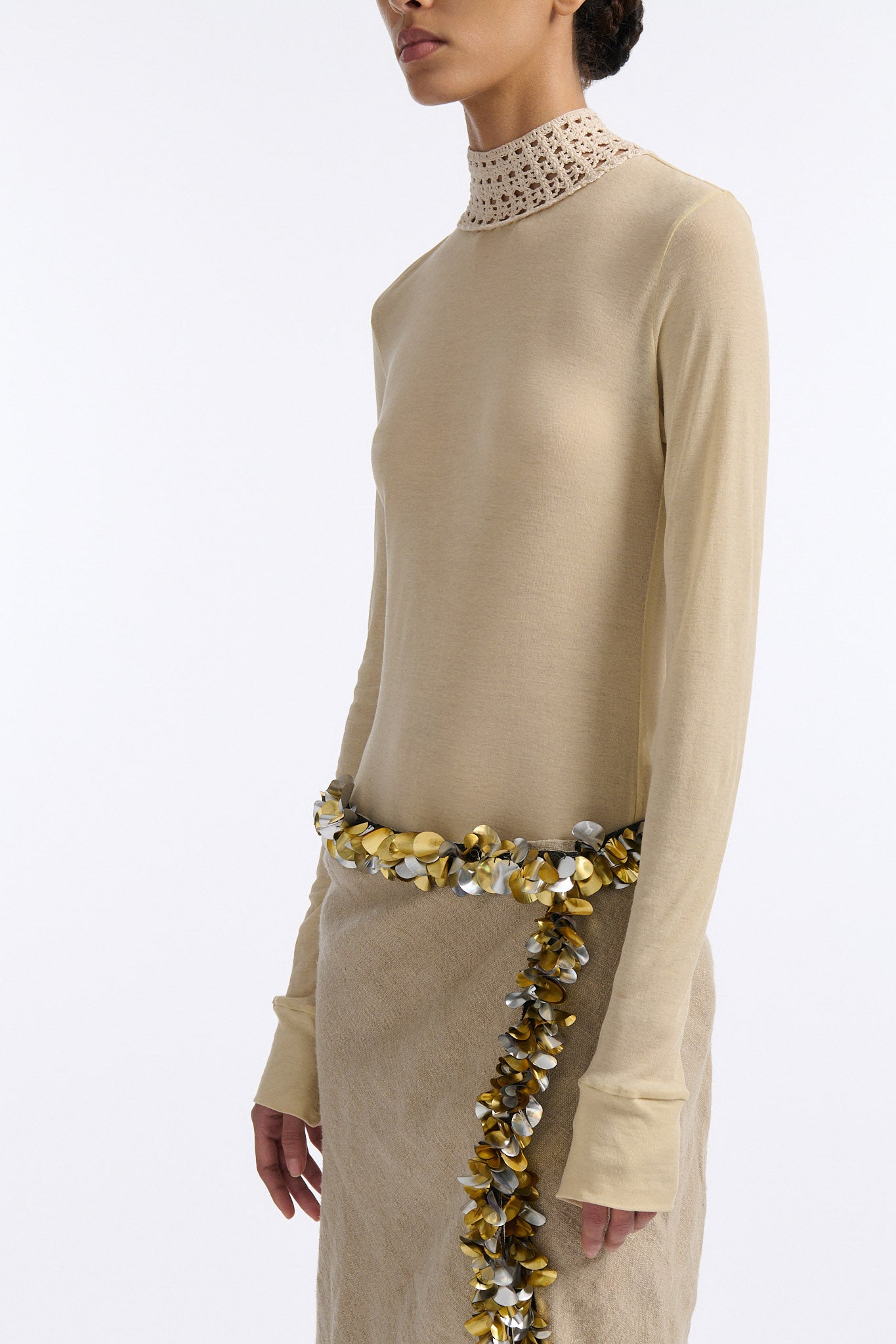 Female model with slicked back black hair in a bun wears beige linen handkerchief wrap skirt. Waist and closure are lined with melted metallic embellishments in gold and silver to provide an oyster illusion. Model also wears light beige fine-gauge cotton long sleeve top with mockneck crochet collar.