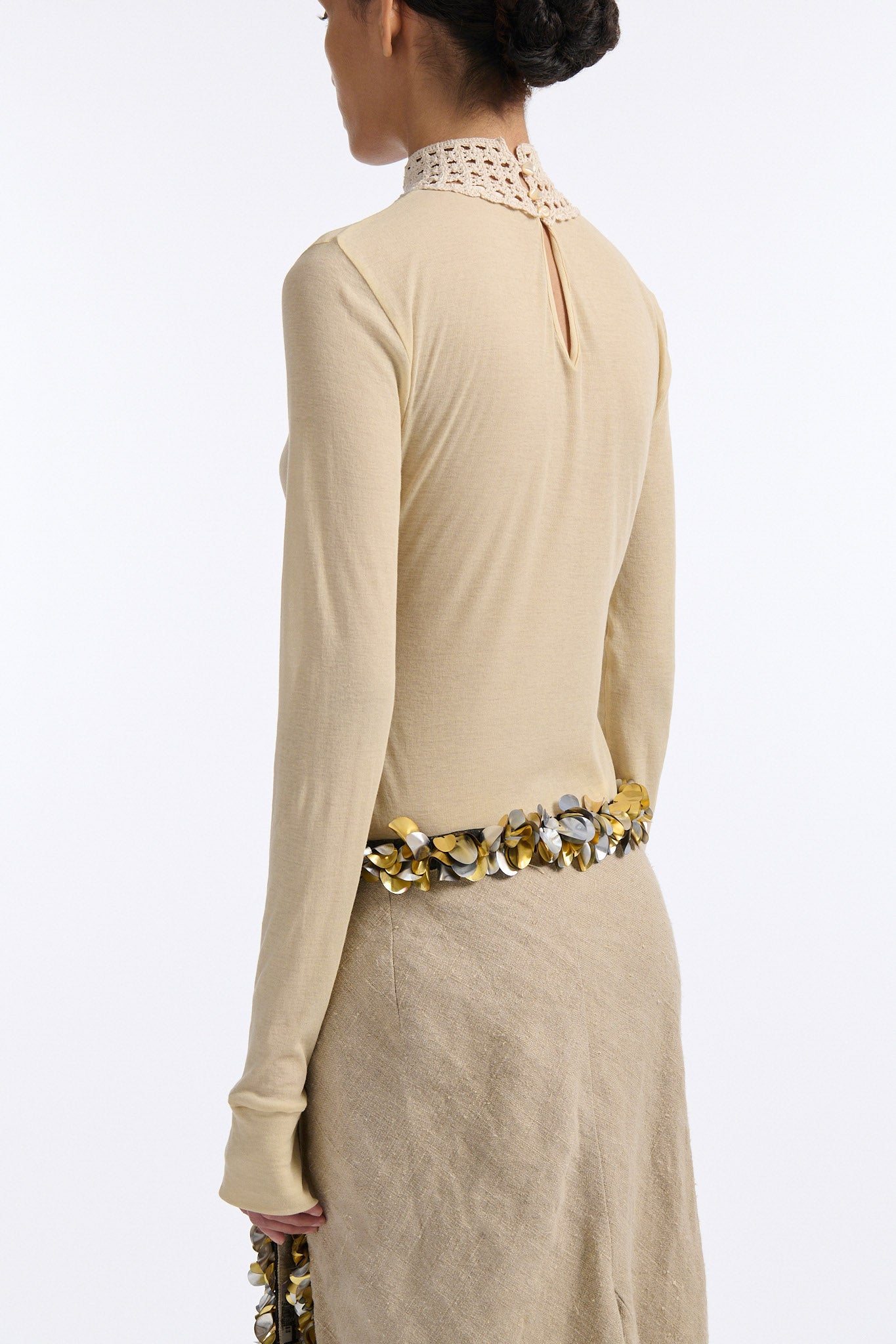 Female model with slicked back black hair in a bun wears beige linen handkerchief wrap skirt. Waist and closure are lined with melted metallic embellishments in gold and silver to provide an oyster illusion. Model also wears light beige fine-gauge cotton long sleeve top with mockneck crochet collar.